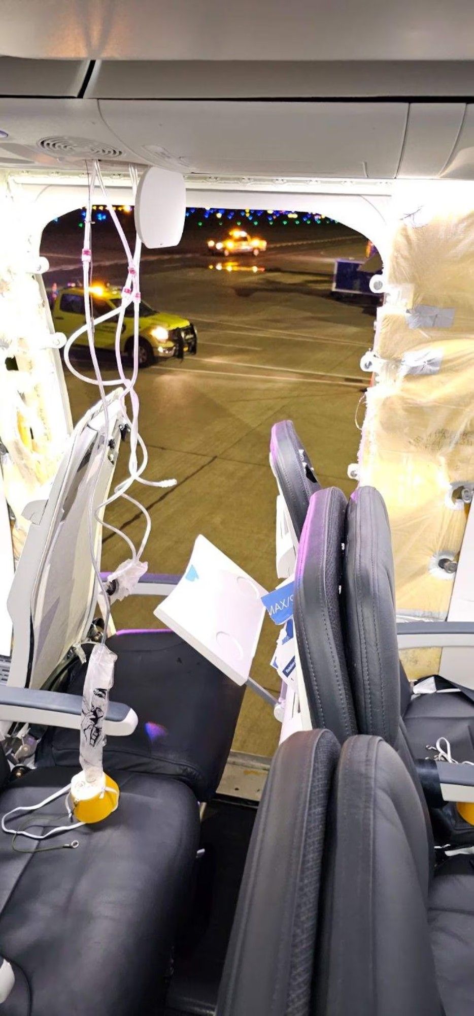 A photo of a seat in an Alaska Airlines 737 Max showing all the black padding ripped off, there's a huge but neat hole in the plane showing a car outside parked on the tarmac. The oxygen masks are deployed.