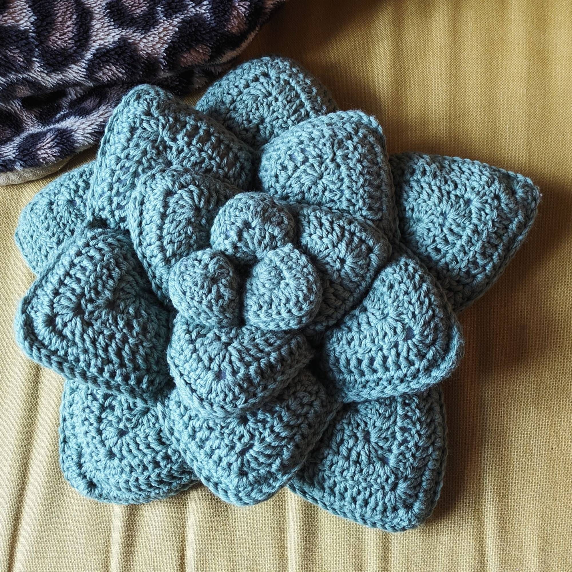 Crocheted Pillow on the shape and greenish-blue color of a succulent. 4 rows of leafs in a starry pattern almost like a flower