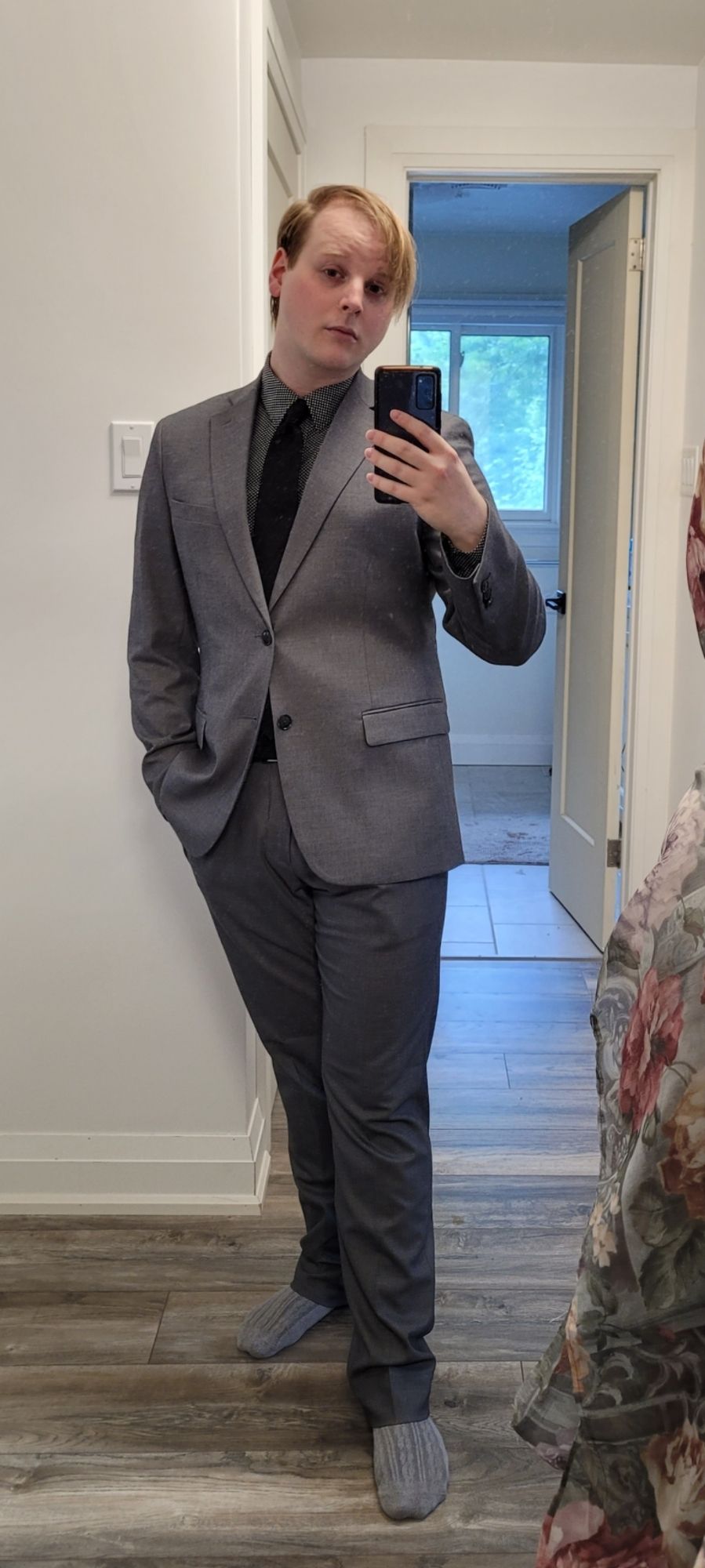 Masculine presenting person in a grey suit, black tie in a hallway mirror.