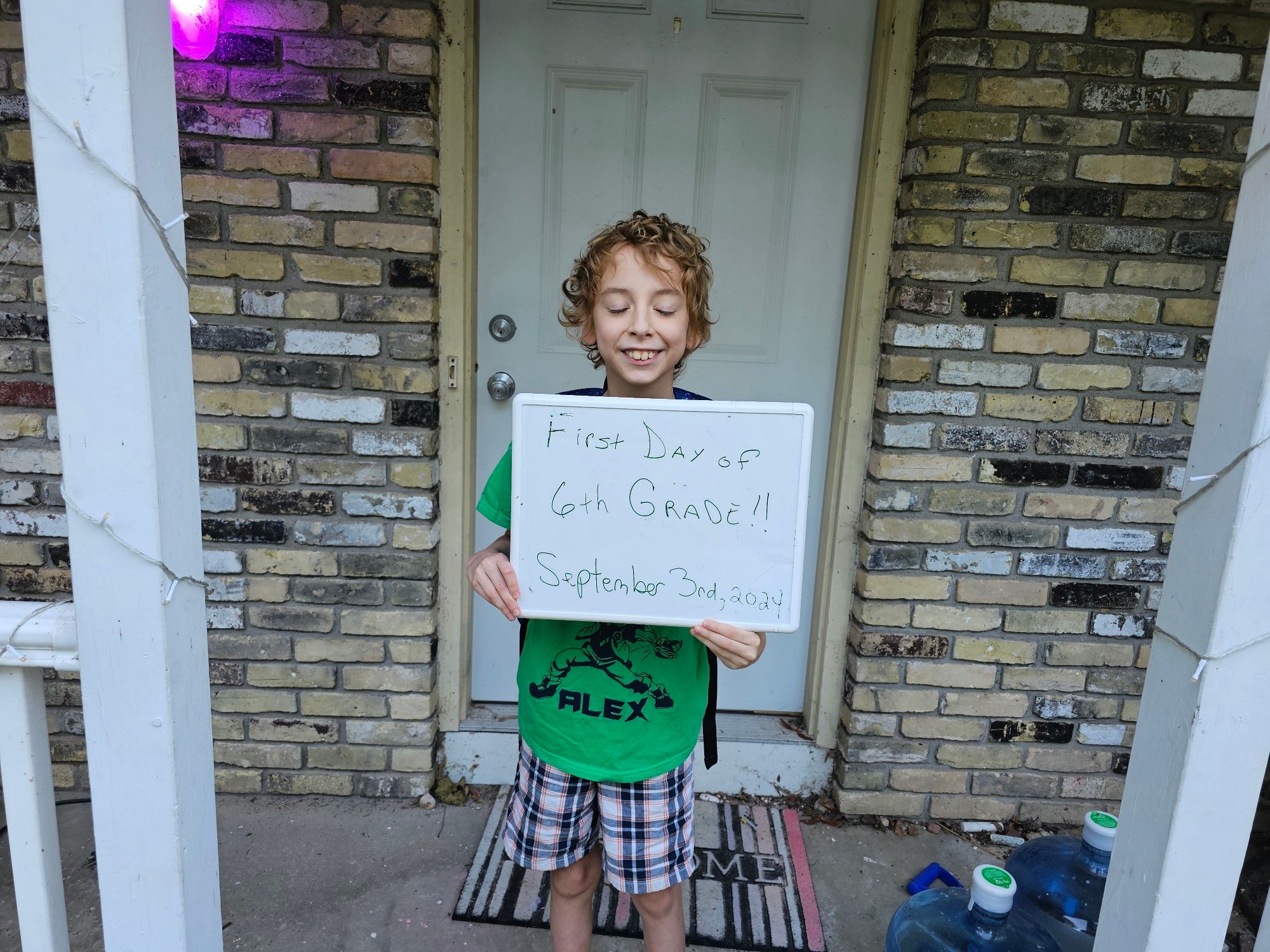 A child in a green shirt holding a sign declaring it his first day of 6th grade.