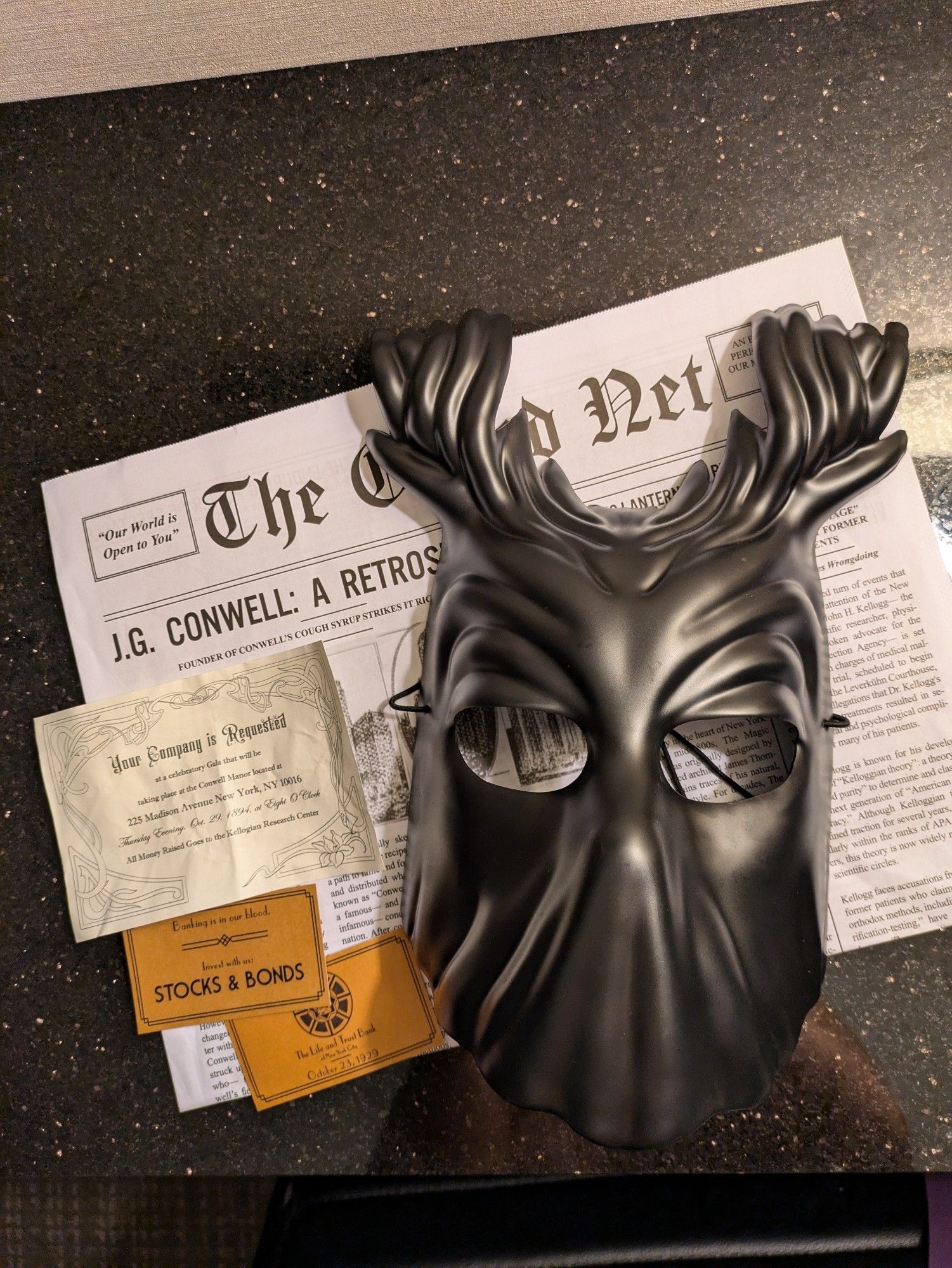 Ephemera from Life & Trust - a creepy black carnivale mask, a newspaper, a weathered gala invitation, bank cards for stocks and bonds