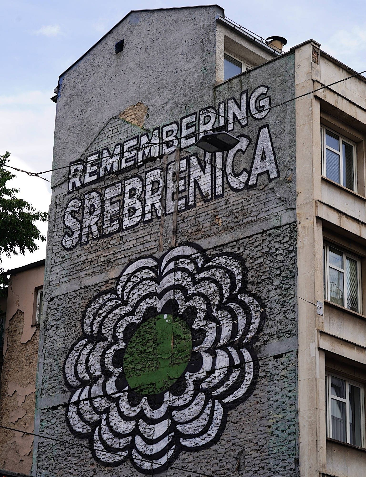 Wandbemalung auf einem alten Haus. Weißer Schriftzug "Remembering Srebrenica " und die ikonische weiß-grüne Blüte.