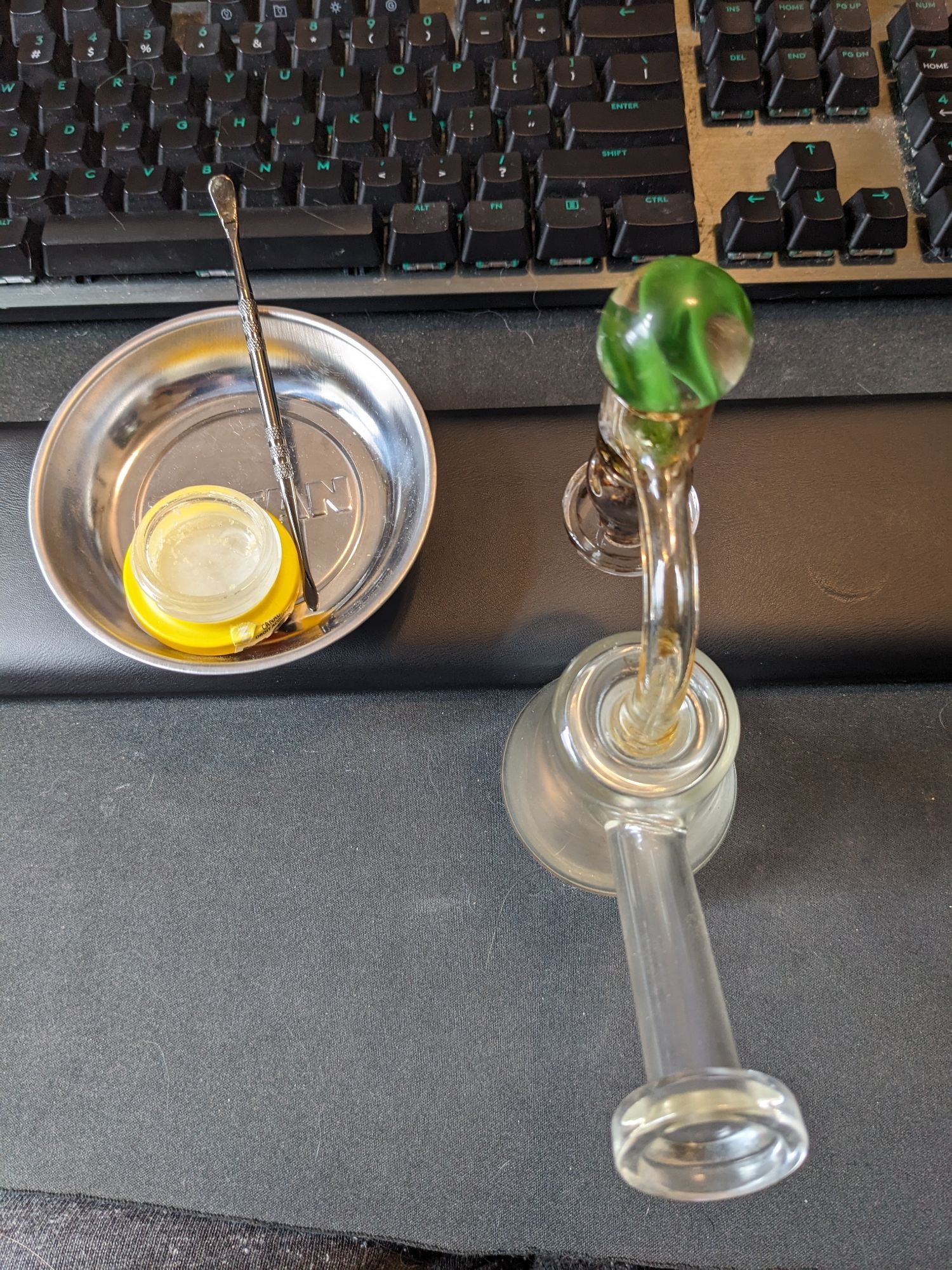 A Titan magnetic screw tray with an open nearly empty hash rosin jar and a metal dad tool sitting in it. The screw tray is holding on to the wrist rest from a Logitech g series keyboard. Above it the keyboard is seen and next to the tray in front of the wrist rest it's a small dab rig with a three marble terp slurper. The marbles are accented with green Glass.