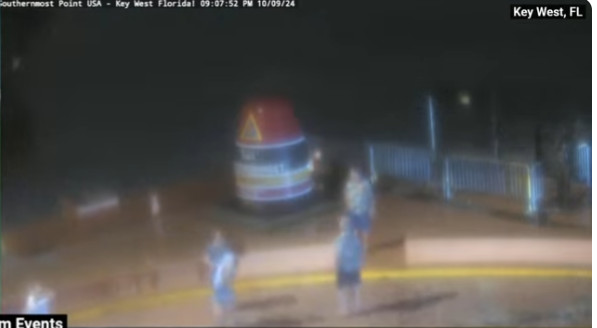 Webcam footage from above of the seafront at night. A large object/landmark in the centre with four people dressed in blue anoraks wondering around 