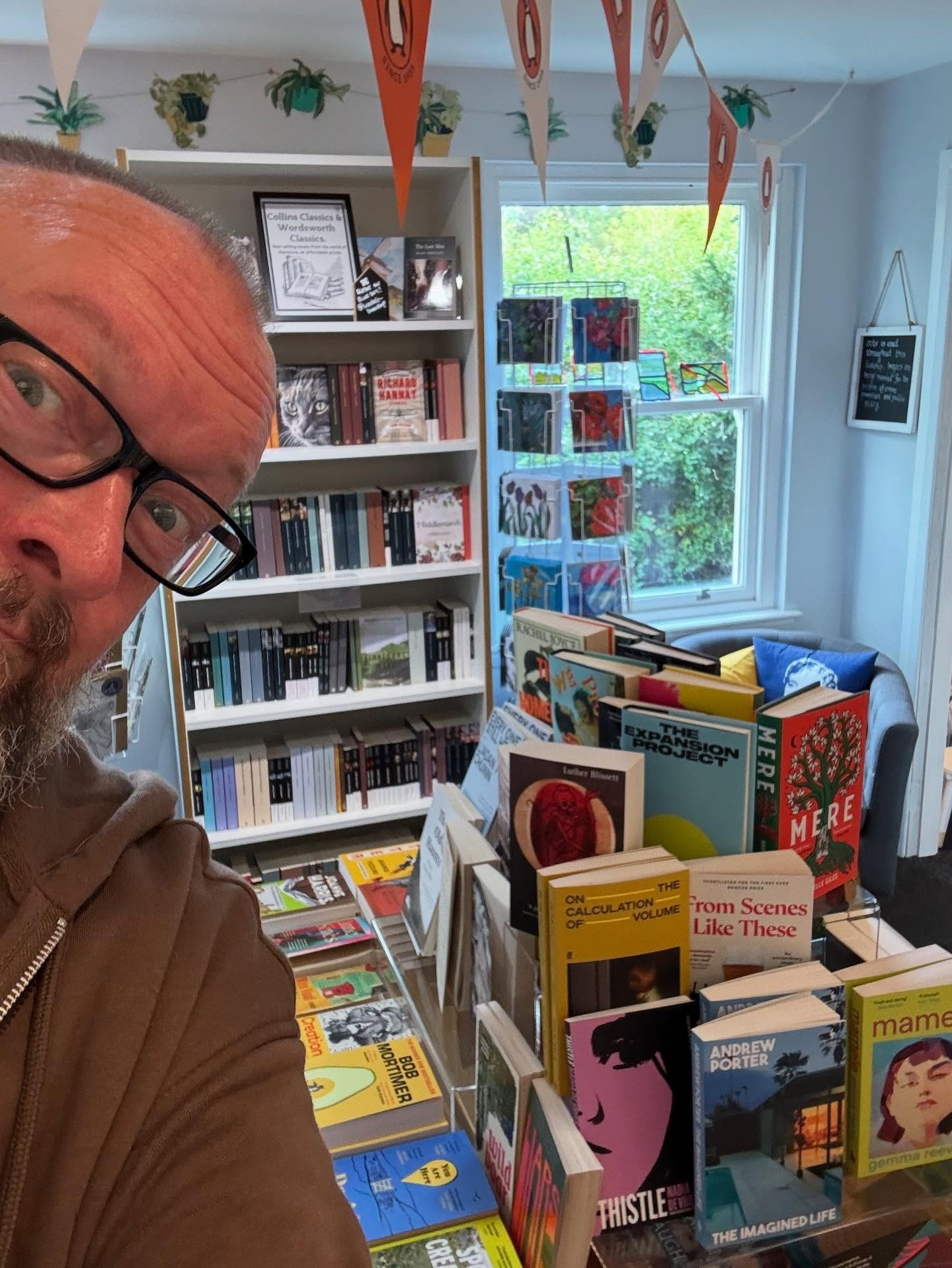 Middle-aged bloke wearing glasses peers around the corner of the image, behind him are bookshelves and a table full of books. 