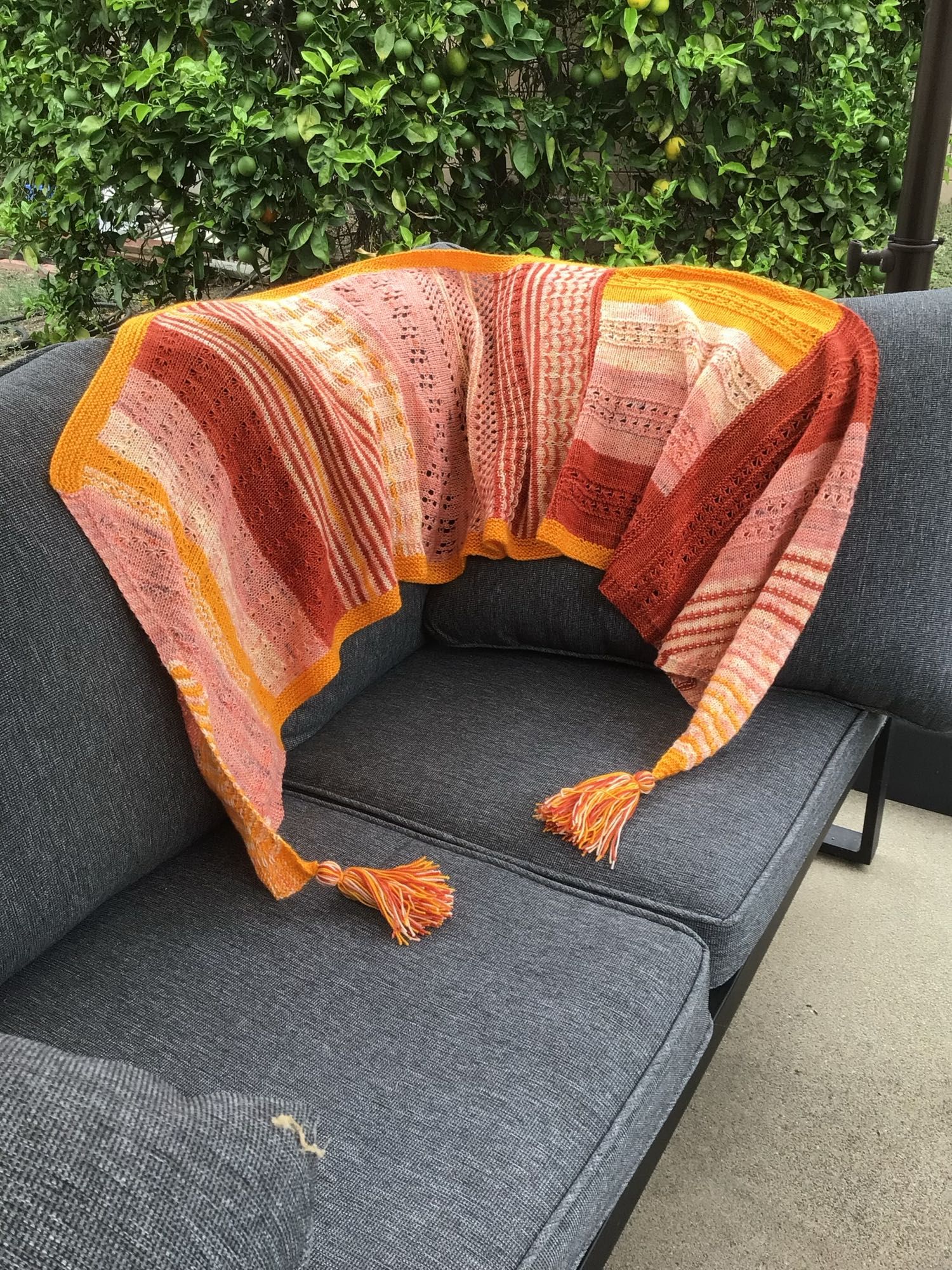 Scarf displayed on a gray couch. Scarf on s multi-colored with peach, rust, ivory, and gold yarns