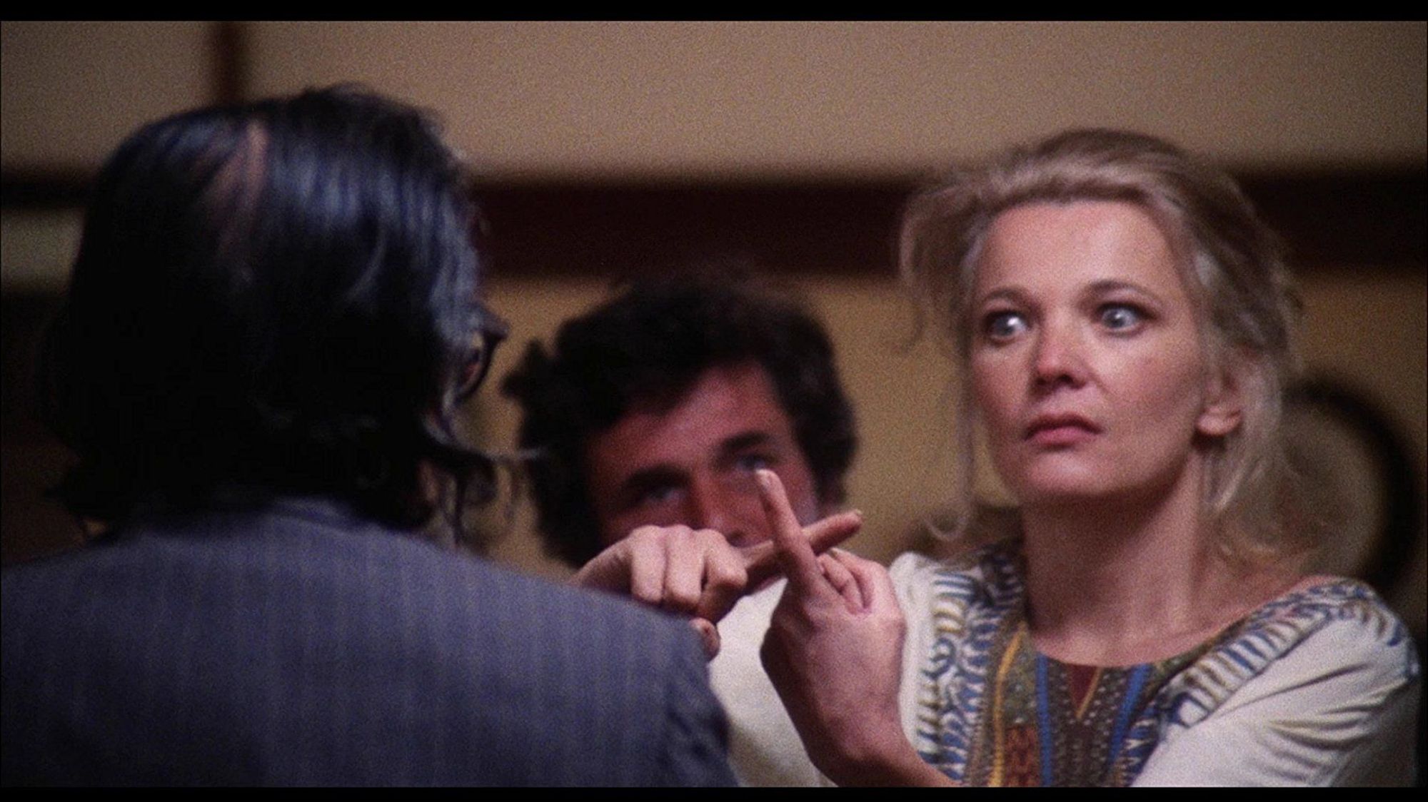 A blonde woman (Gena Rowlands) with a crazed look in her eyes holds her fingers in the shape of a cross at a houseguest in a suit with greasy hair, as her bewildered husband (Peter Falk) looks on from behind her.