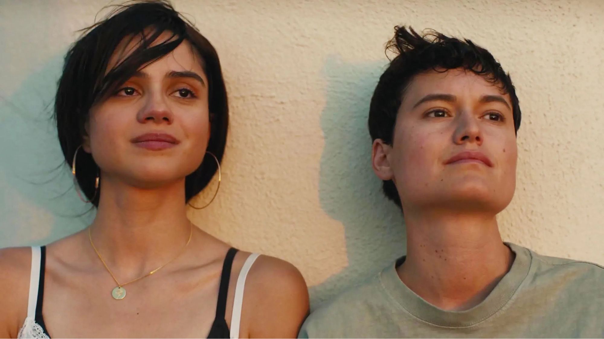 Two Latina sisters (Sasha Calle and Lio Mehiel) stand against a wall at the airport looking wistfully at the sunset after a yearly visit with their deadbeat alcoholic dad.