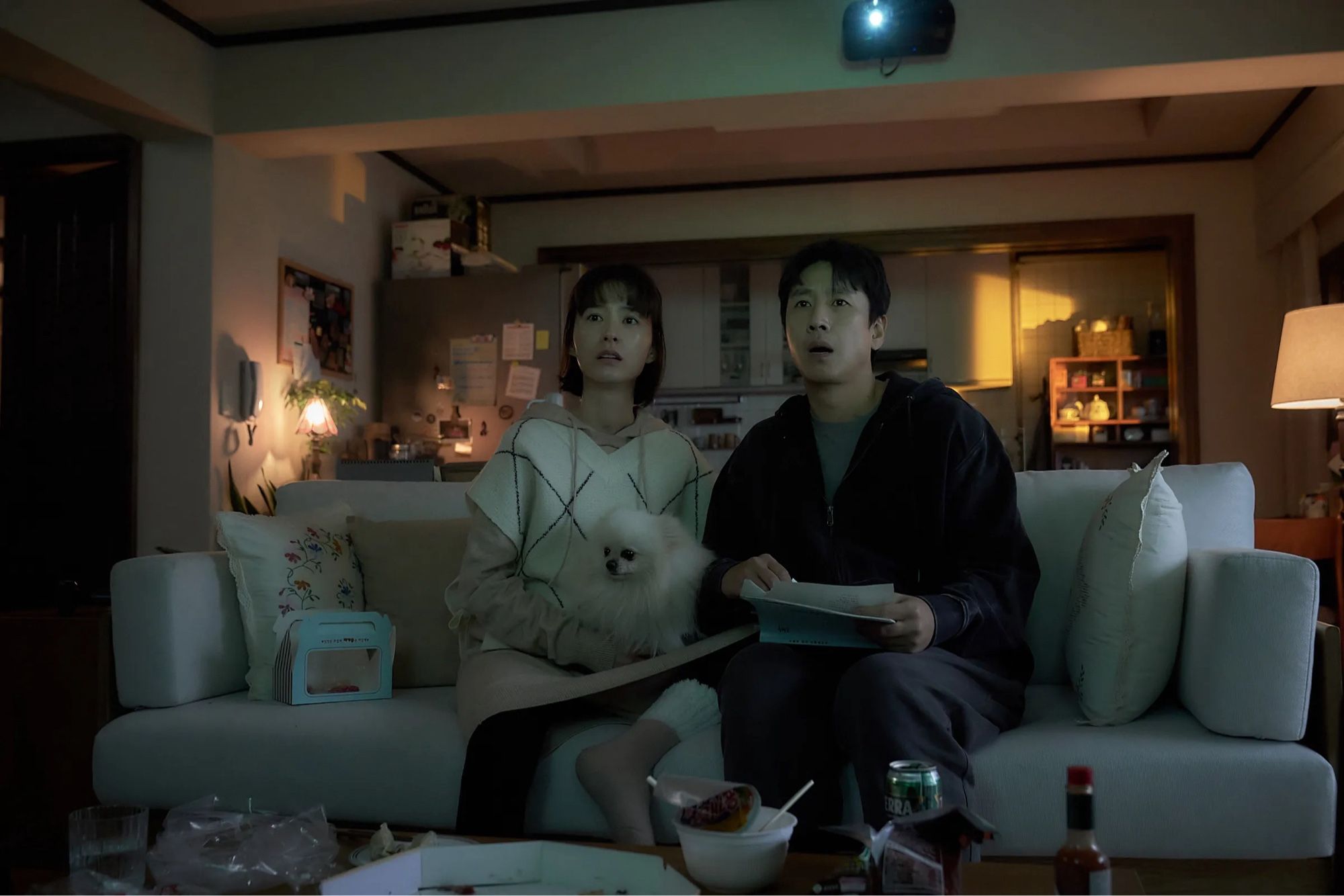 A young Korean couple expecting a baby sit on a couch with their dog watching TV on a projector