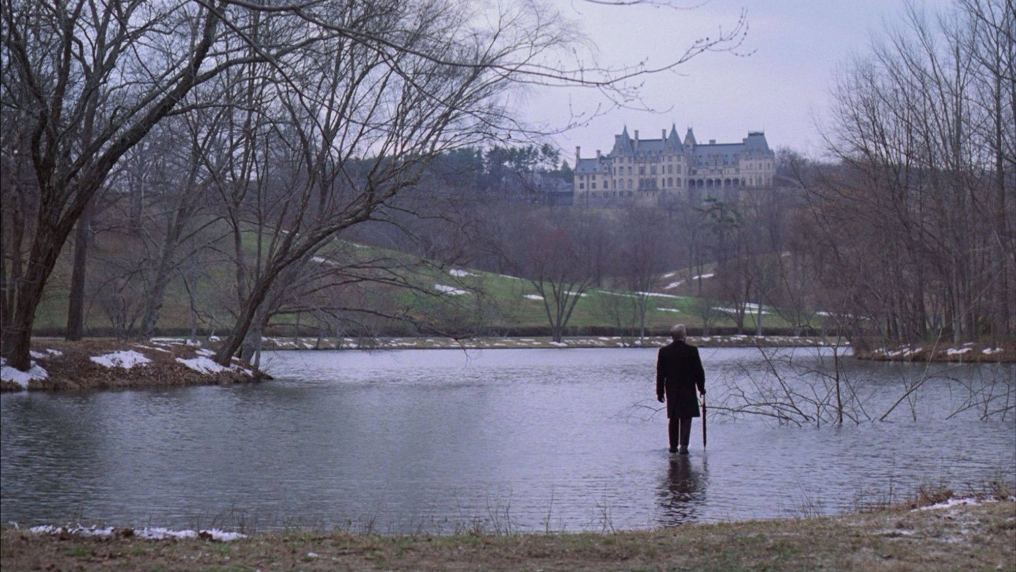 A man in a suit (Peter Sellers) walks on water across a lake towards a mansion in the distance.