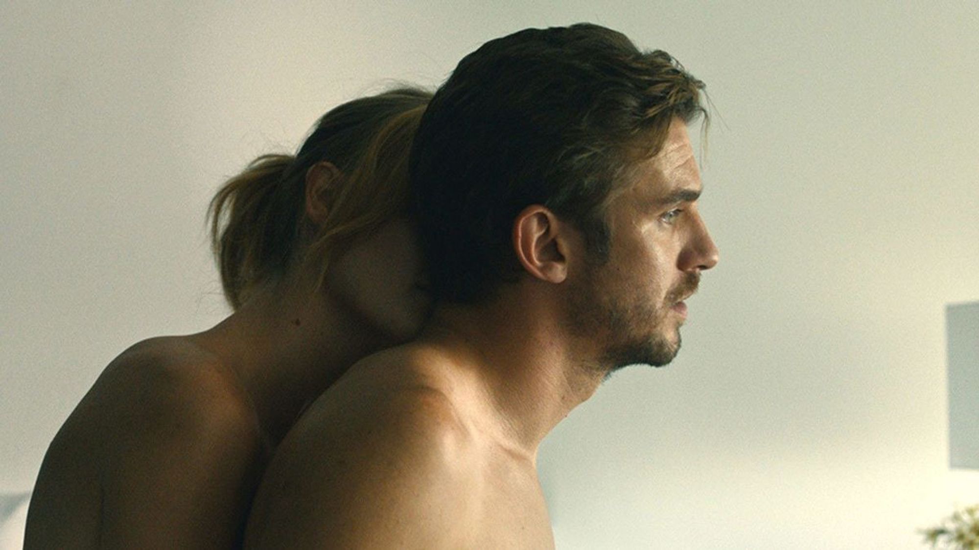 A woman (Kerry Bishé) kisses the back of the neck of the man she's sleeping with (Dan Stevens), as he looks offscreen .