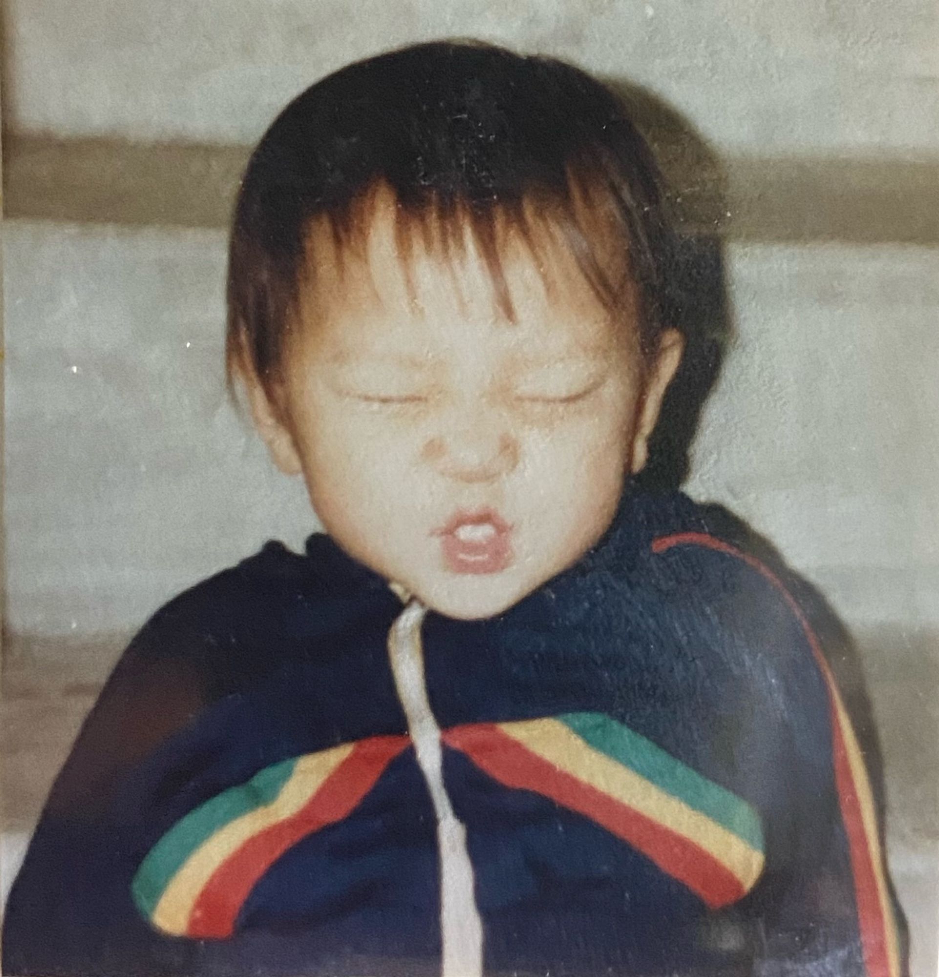 A korean baby with wispy, dark hair ina navy hoodie with rainbow stripes squeezes their whole face shut in an attempt to smile