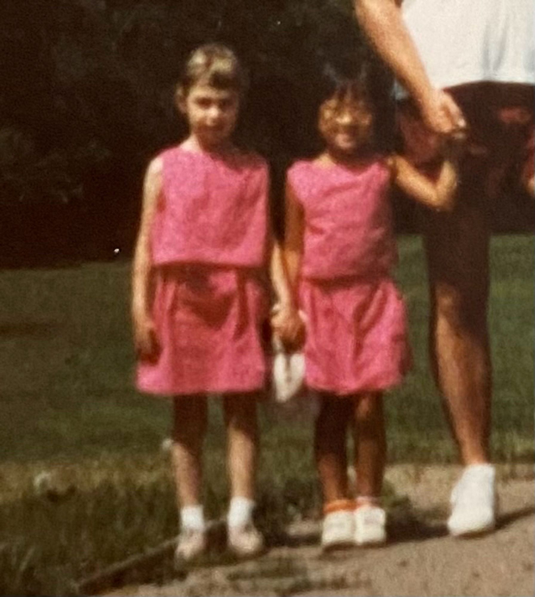 A blurry phot of two little kids dressed identically in pink tops and shorts, the white girl on the left is pale and blonde, the east asian kid on the right is tan and wearing glasses, arm holding on to the hand of a tan adult mostly cut out of the photo