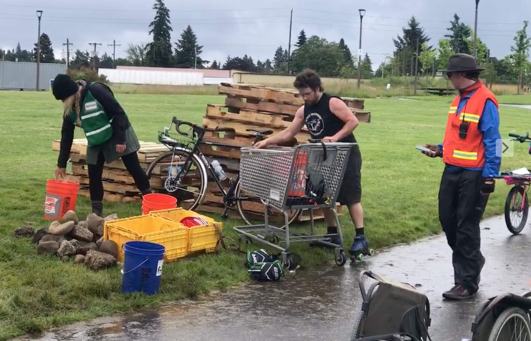 3 people, two in vests acting as officials for the Portland DRT (Disaster Relief Trials). Around them are stacks of pallets, piles of stones, and other materials which were items in the tests of the DRT. In the center of the photo, Ryan Hashagen, with a shopping cart. They are wearing shorts, a sleeveless tshirt, and roller skates, which is their usual garb for most of the year.