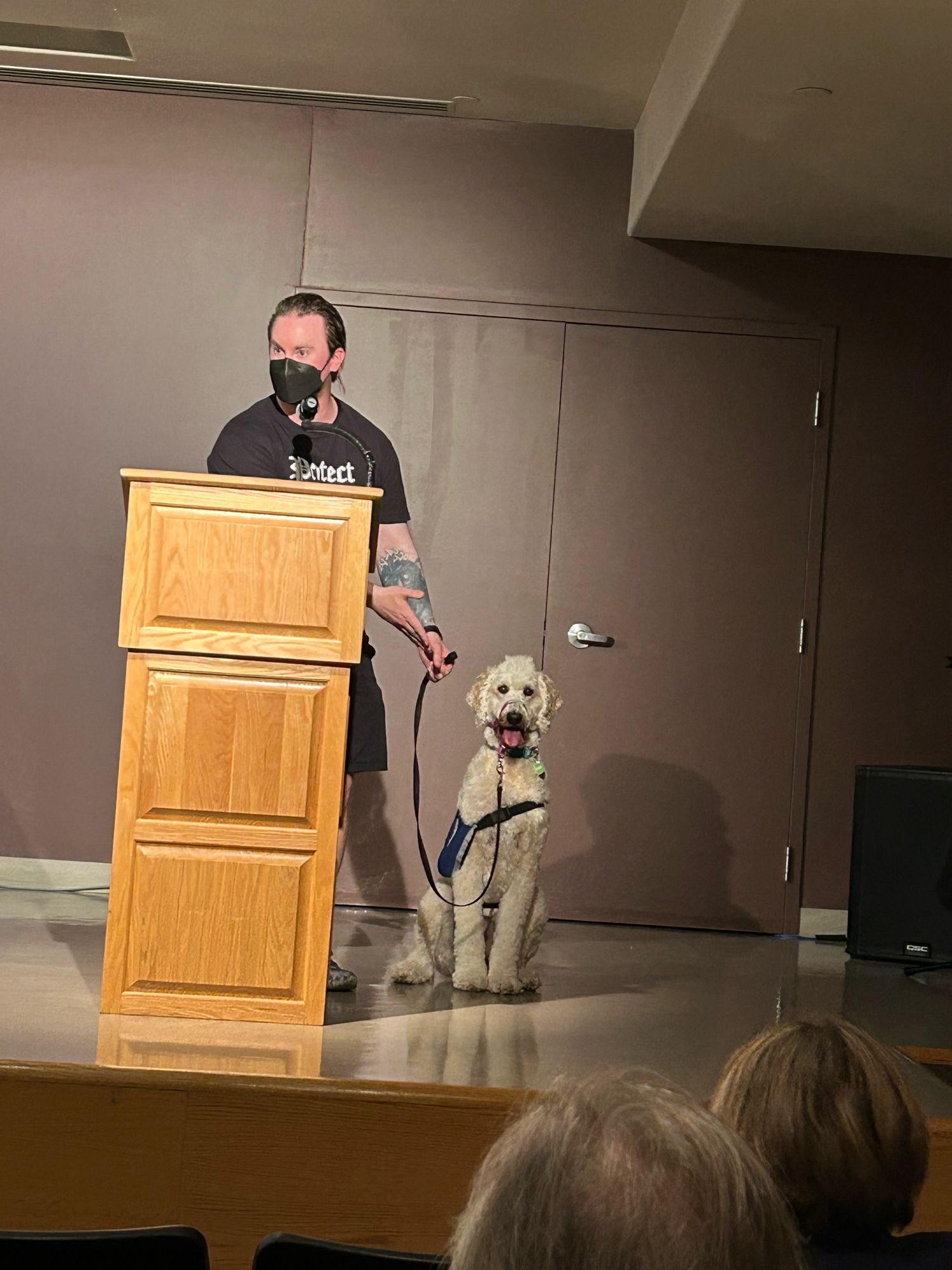 View from within an audience as a large white goldendoodle sits politely beside a bearded white man in an N95 mask. The man stands behind a podium. Audience members watch them.