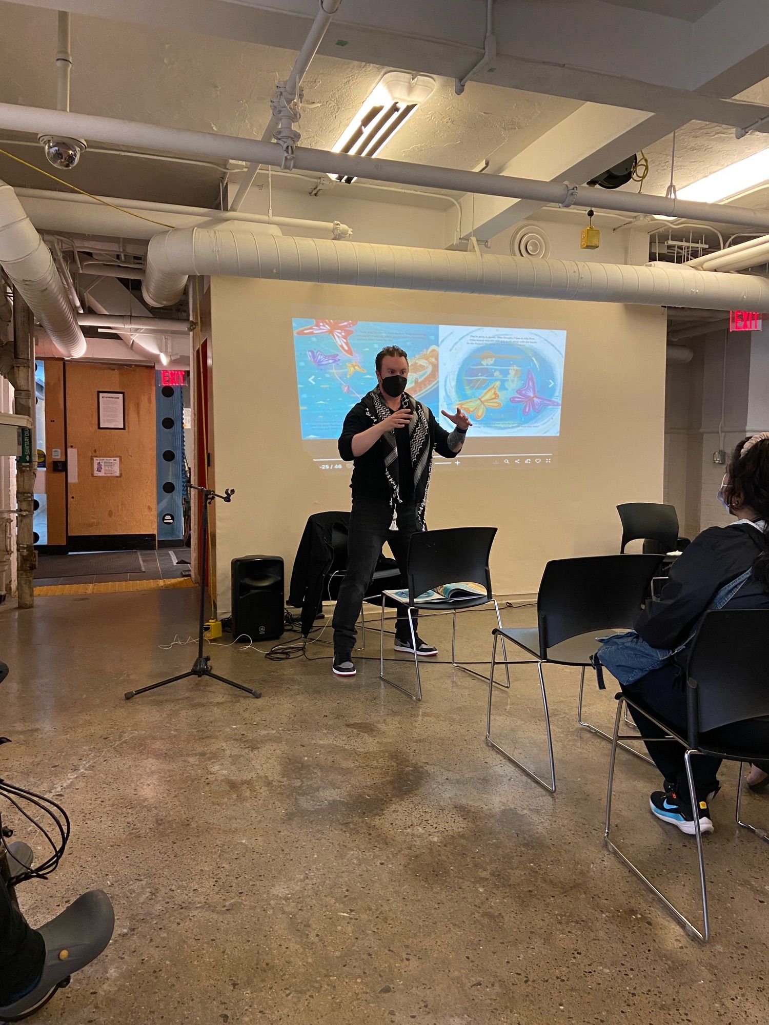 Author Adam Pottle performs his children’s story BUTTERFLY ON THE WIND in American Sign Language with the illustrations projected behind him. He wears a black N95 mask, black shirt, black pants, and a black and white keffiyeh.