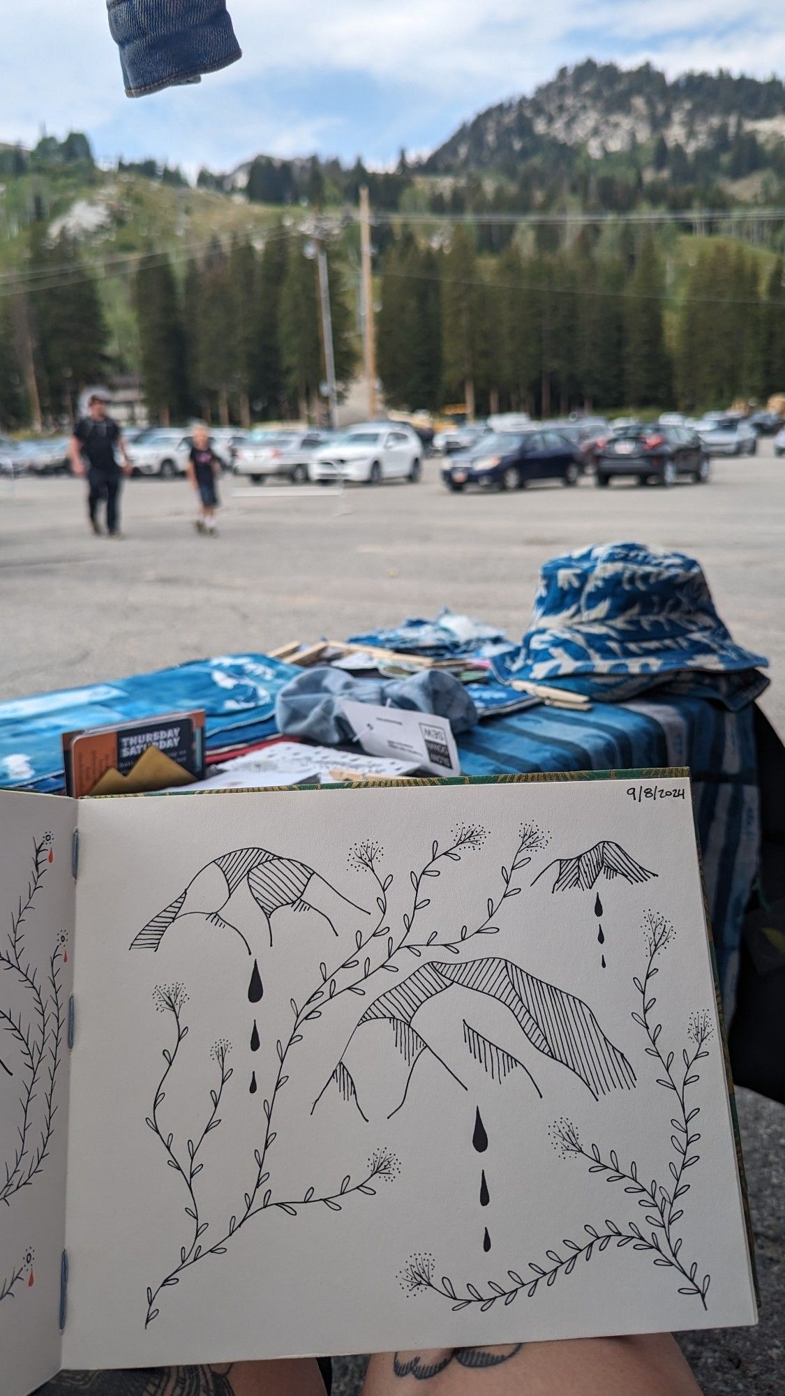 A sketch of 3 mountains with 3 tears under each and simple plants weaving around them. In a parking lot with a view of mountains from the artists pov sitting in their booth.