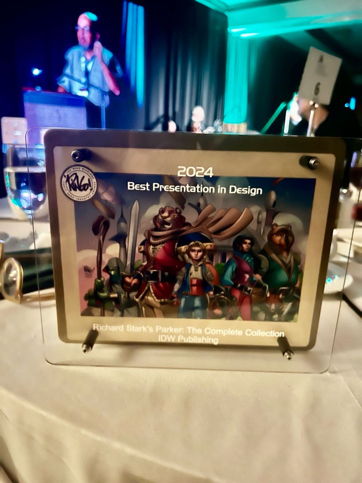 An award sits on a table, it is a Ringo Award for Best Presention in Design for Richard Stark’s Parker: The Complete Collection, IDW publishing. 

The award is rectangular, and has a background of cartoon characters, and has a raised glass cover above.