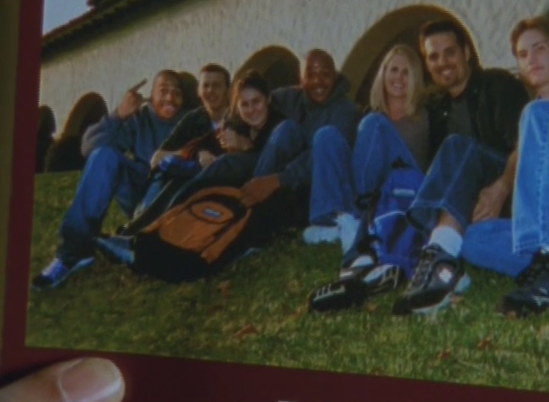 A hand holding a brochure with a picture of smiling college students sitting togther on a lawn. All the students are white except two, who are actually the same young bald black man in a hoodie photoshopped to be in two places at once