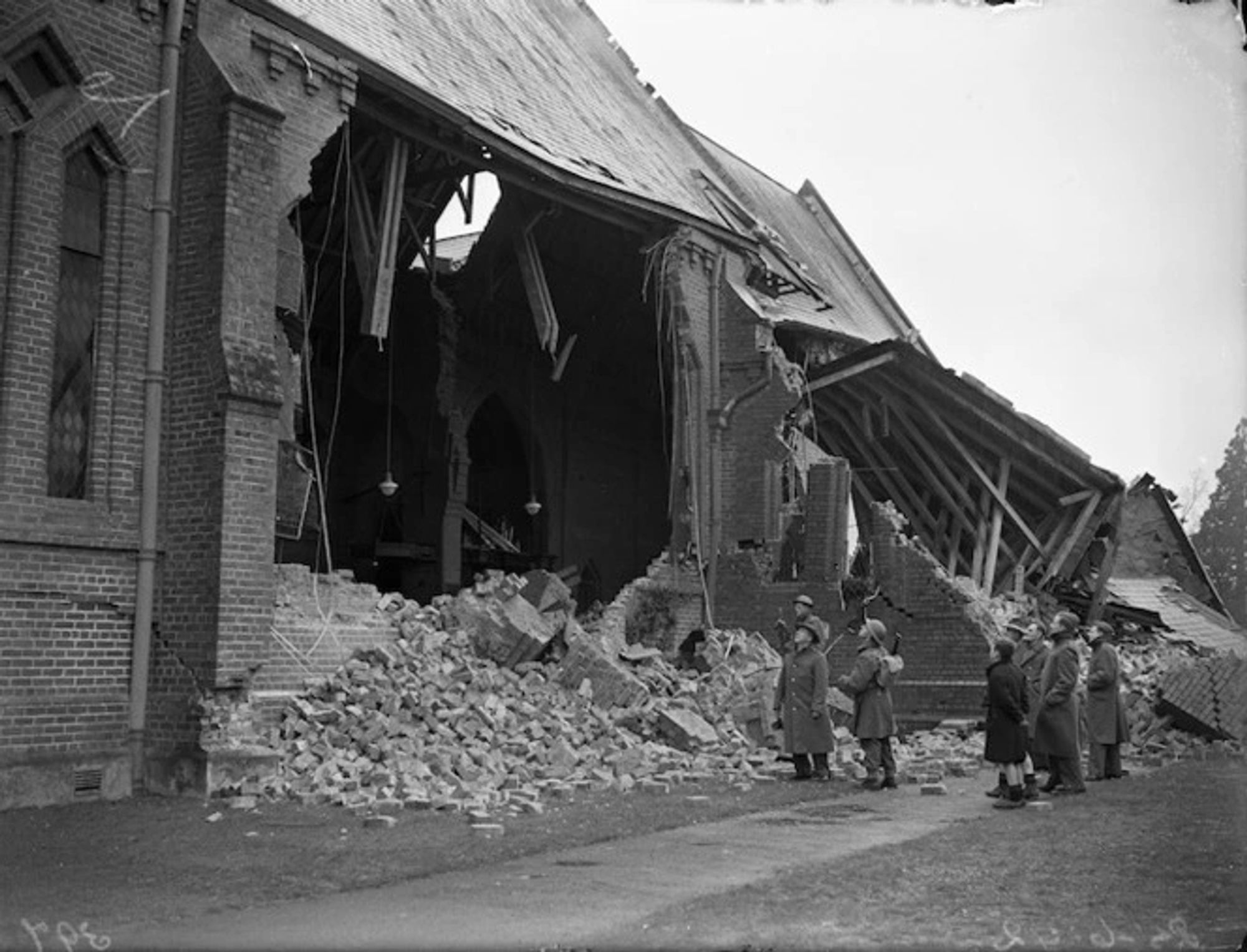 A black and white photo of people looking at a damaged brick church. Walls have collapsed and the roof hangs unsupported.