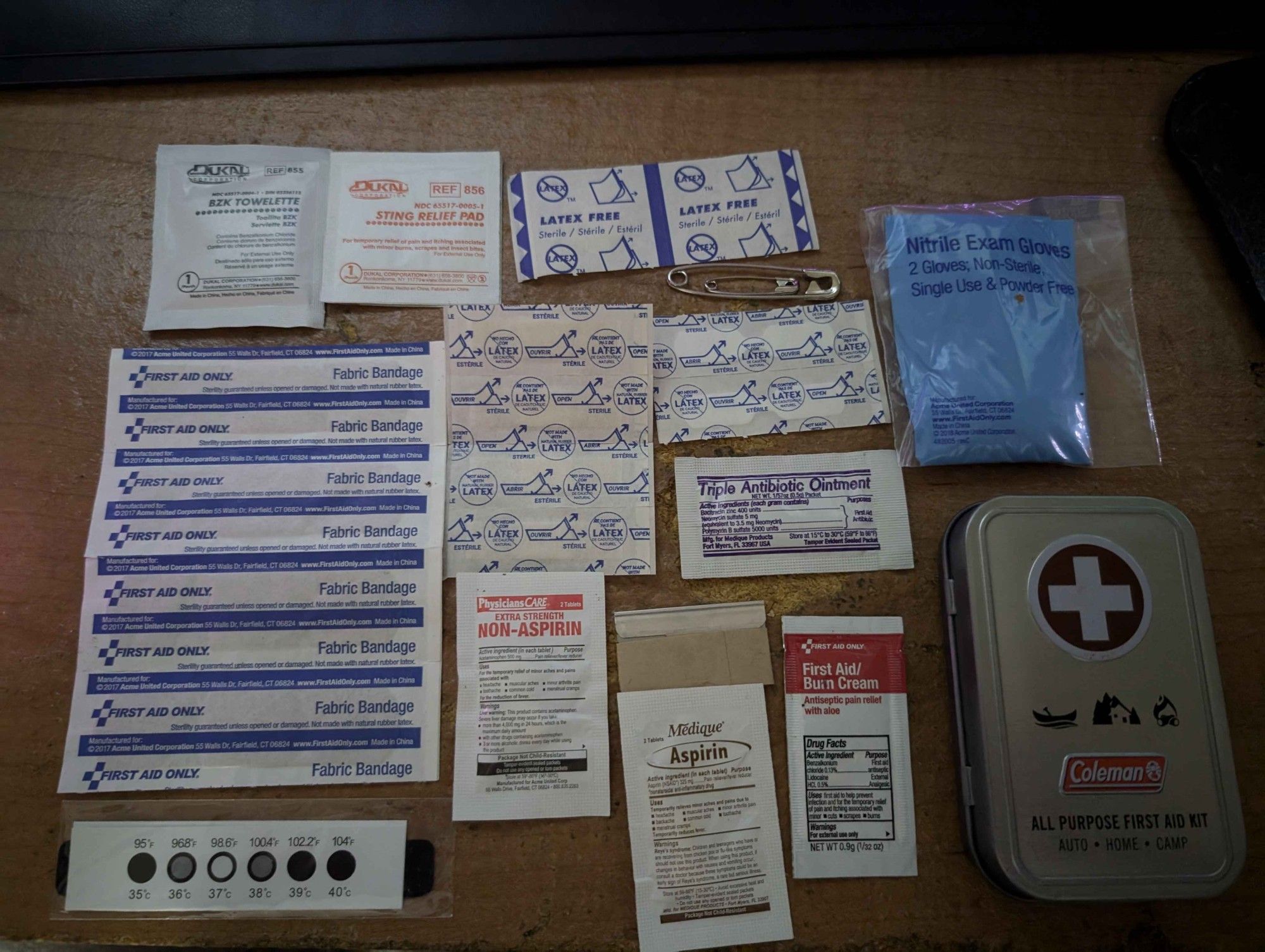 The contents of a Pocket First Aid Kit on display:
BZK Towelette
Sting Pads
2x spot bandages
2x safety pins
Pair of non-sterile nitrile exam gloves
A long strip of 4 fabric bandages
A smaller strip of 4 small bandages
2 butterfly closures
Triple Antibiotic Ointment
A fever strip
Non-Aspirin Pain Relief
A razor blade in cardboard
Aspirin
Burn Cream
A Coleman All Purpose First Aid Kit Metal Tin