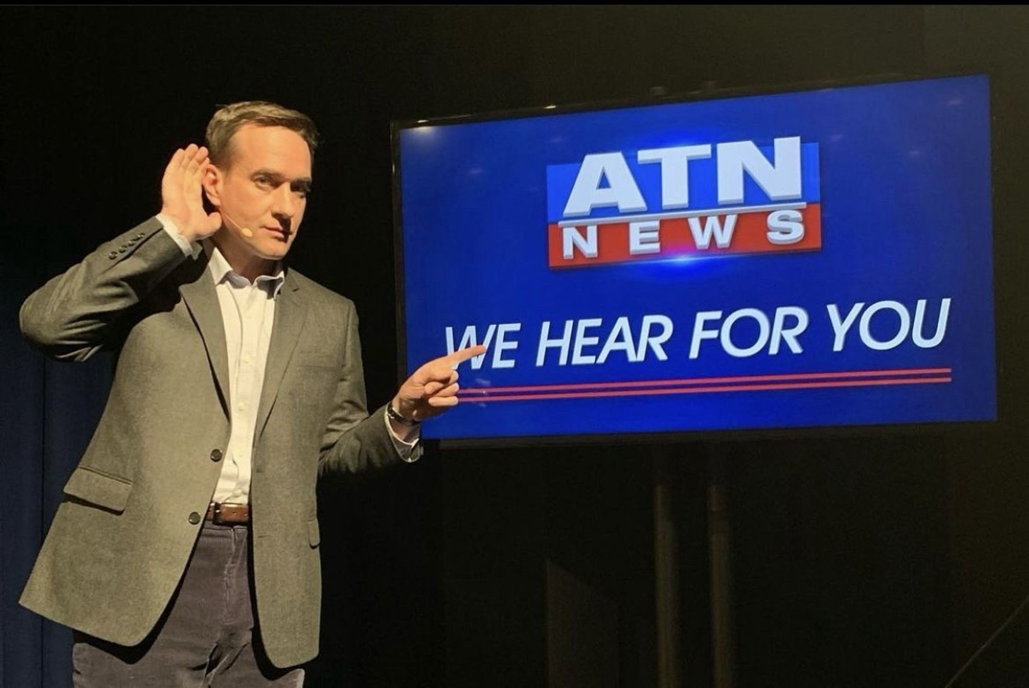 Matthew Macfadyen poses as the character Tom Wambsgans from the television series Succession in front of a screen displaying the fictional ATN News network’s slogan, “We hear for you.”