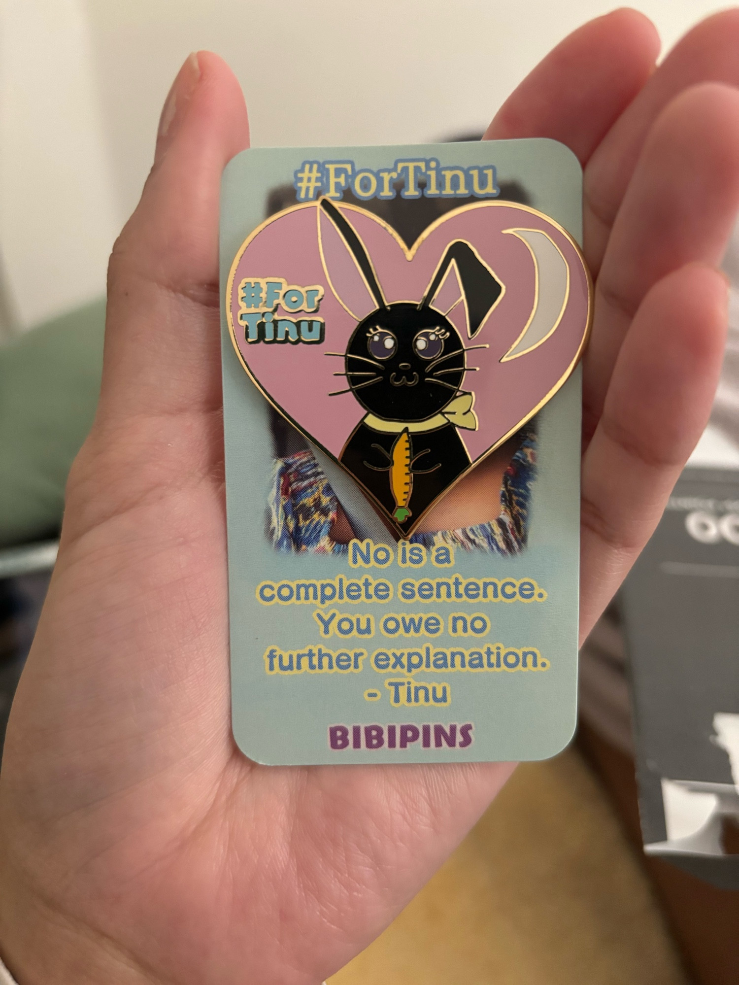 fortinu pin by Bibi Pins. The words #fortinu are in the corner. A black rabbit with purple/pinkish fur on the inside of its ears wearing a cute yellow neck tie/scarf holding a carrot. A white crescent moon is in the corner and all of this is inside a golden rimmed pink heart which is the shape of the pin. The text on the card is beneath a picture of Tinu and says “no is a complete sentence. You owe no further explanation. - Tinu” BIBIPINS