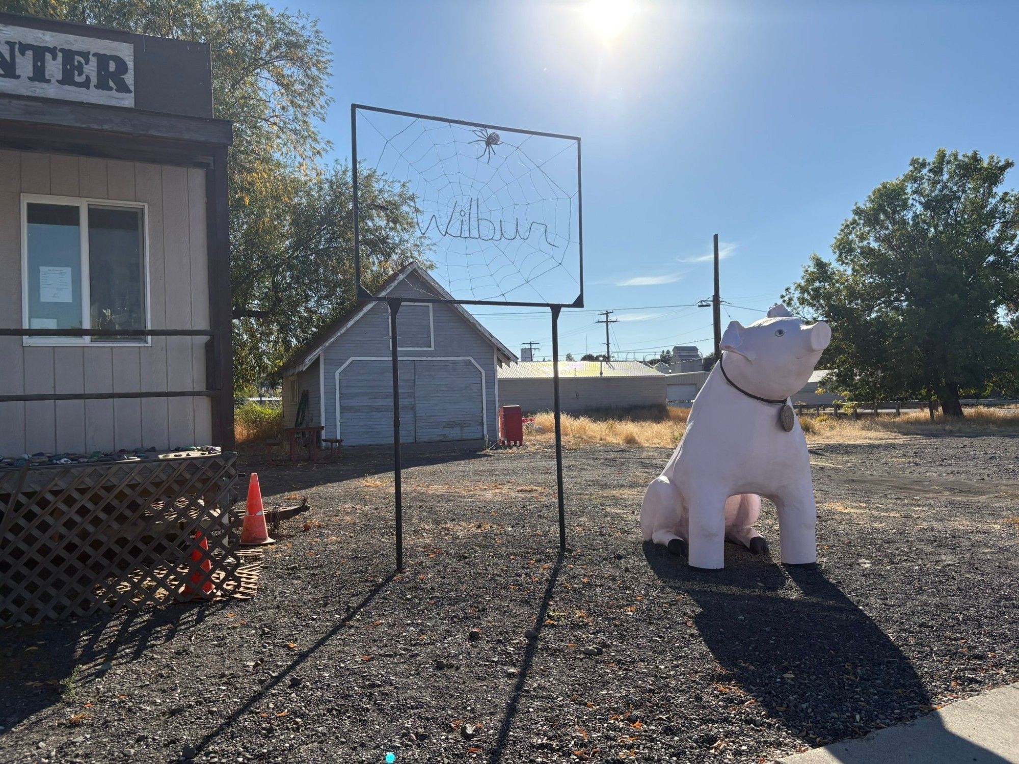 a fiberglass (?) statue of a pink pig, with a collar and a name tag. It is on gravel, in front of a visitor center for a small town, and in the background is a wire spiderweb, with a spider, and the word in the web is, "Wilbur" which is the name of the pig in Charlotte's Web.