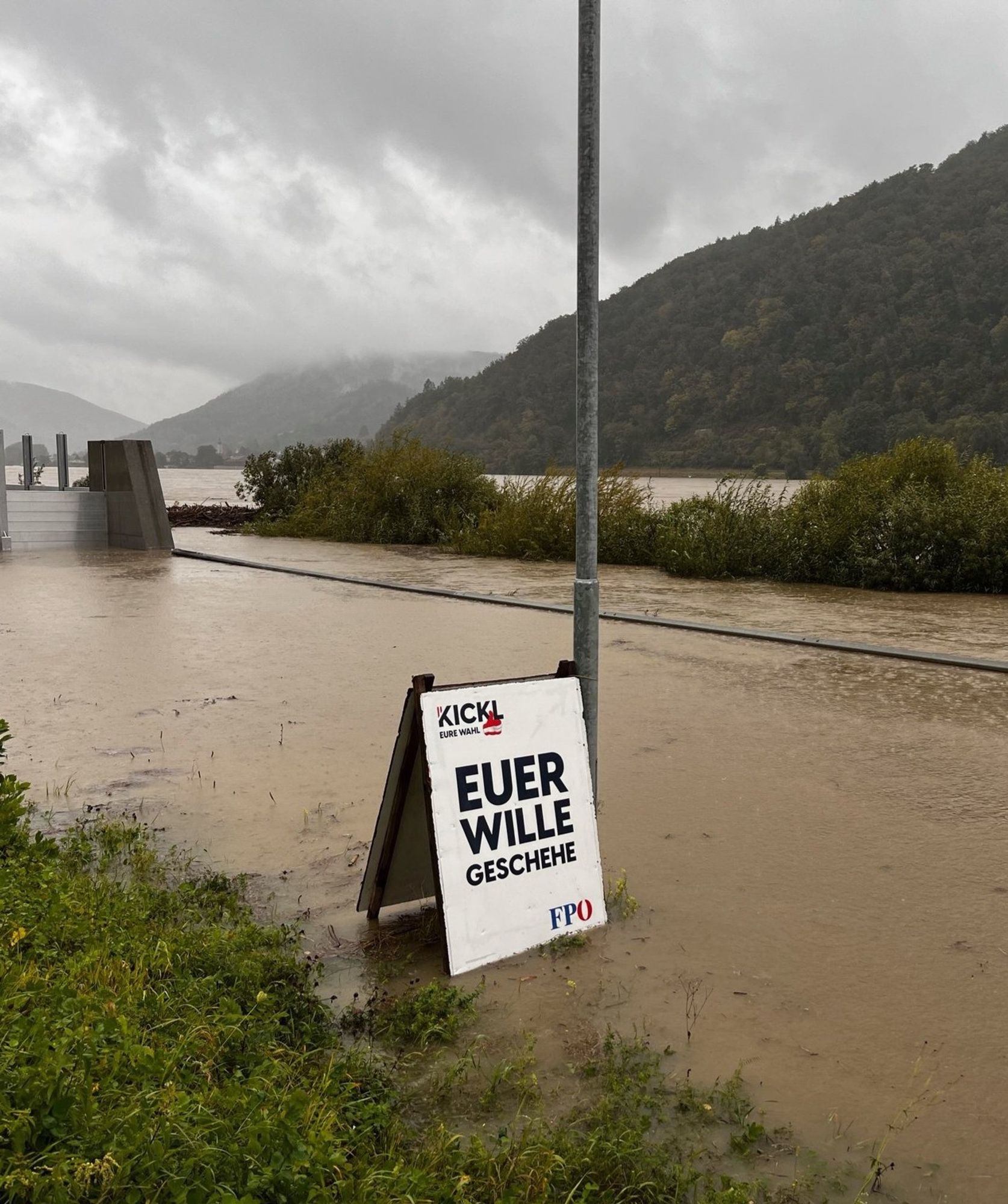 Ein Wahlplakat der FPO steht auf einer überfluteten Straße, neben einem Fluss. Im Hintergrund hügelige Landschaft mit Wäldern. 
Text auf dem Plakat: Euer Wille geschehe 
Kickl Eure Wahl