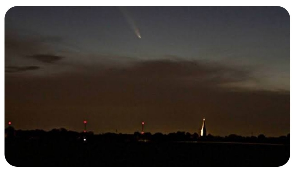 Am dunklen Himmel ist ein Schweif erkennbar. Im unteren Teil des Bildes sind kleine Lichter und ein angestahlter Kirchturm 