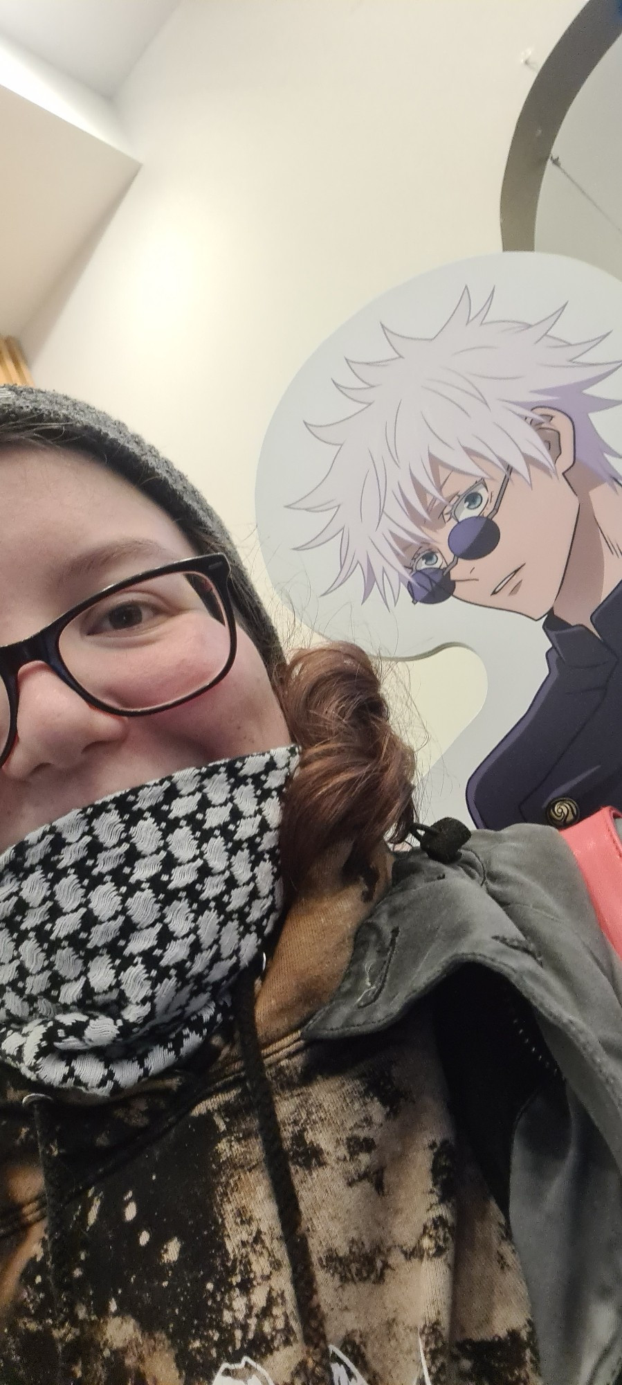 Selfie einer Frau mit Beanie. Sie lächelt glücklich. Im Hintergrund sieht man einen Pappeaufsteller der Figur Gojo Satoru aus dem Anime Jujutsu Kaisen.