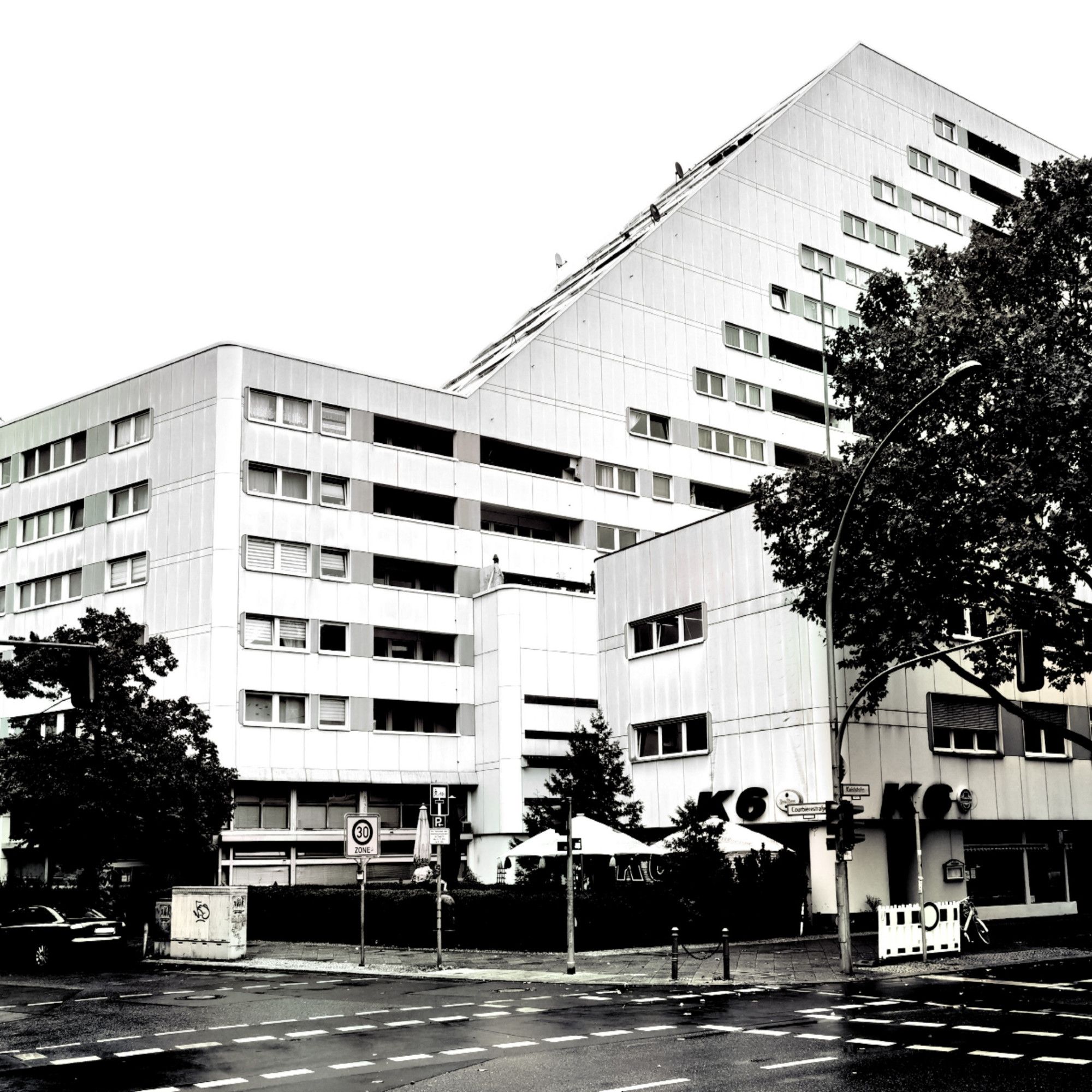 Das schwarz-weiße Foto zeigt die Kreuzung der Courbièrestraße mit der Kleiststraße in Berlin-Schöneberg. Das Haus, in dem Irene von Orlow lebte, steht schon seit langer Zeit nicht mehr. Der Blick auf dem Bild führt über die Kreuzung und zu einem kleinen Biergarten hinter einer akkurat gepflegten Hecke an der Straßenecke. Baumkronen zweier großer Laubbäume befinden sich an beiden Bildrändern, im Bildhintergrund erhebt sich ein sehr großer, moderner und mehrgeschossiger Neubaublock, der in Berlin gemeinhin als "Sozialpalast" bezeichnet wird. Die Wohnanlage hat eine helle, größtenteils saubere Fassade mit zahlreichen Fenstern. Der Himmel im oberen Bildbereich ist sehr dicht bewölkt, aber hell.