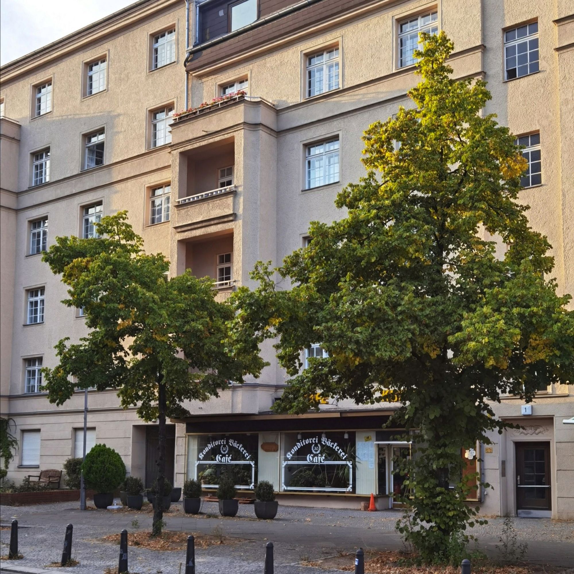 Das Foto zeigt das mehrgeschossige Mietshaus in der Hildegardstraße 1 in Berlin-Wilmersdorf. Der Blick auf dem Bild führt hindurch zwischen zwei großen, üppig grünenden Laubbäumen, die das Haus teilweise verdecken. Die Fassade des Hauses ist beige und sauber und weist zahlreiche, große Fenster und mehrere Balkone auf. Im Erdgeschoss des Hauses befindet sich eine Bäckerei mit kleinem Café.