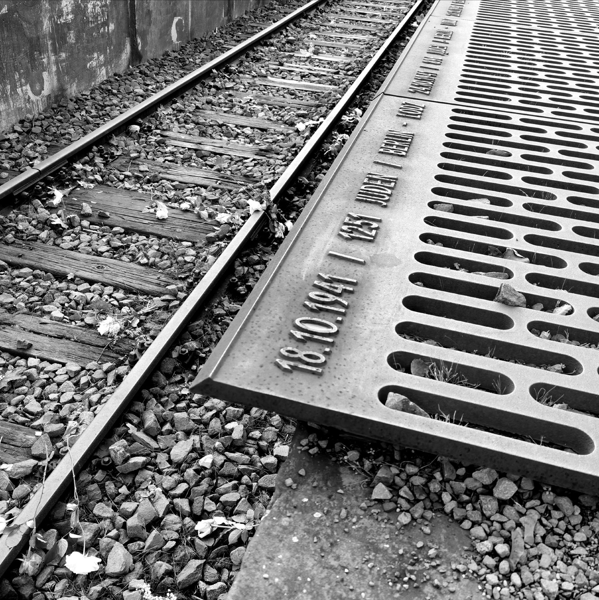 Das schwarz-weiße Foto zeigt am linken Bildrand das Gleis und das Gleisbett des heutigen Mahnmals "Gleis 17" am Bahnhof Grunewald in Berlin. Weiße Rosen liegen auf den beiden Schienensträngen sowie im Schotter dazwischen. Die bronzene Bodenplatte mit dem Datum des ersten Deportationszuges aus Berlin befindet sich am rechten Bildrand, auf ihr die eingravierte Inschrift: "18. 10. 1941 / 1251 Juden / Berlin - Lodz".