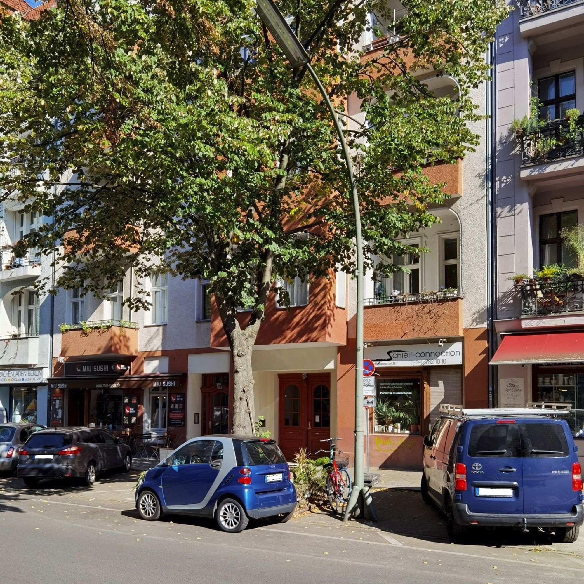 Das Foto zeigt aus einem seitlichen Blickwinkel das mehrgeschossige Mietshaus in der Eisenacher Straße 80 in Berlin-Schöneberg. Der Blick auf dem Bild führt von der Straße hinweg über bzw. vorbei an mehreren Pkw, die vor dem Haus geparkt sind. Das Haus hat eine saubere Fassade mit zahlreichen, großen Fenstern und mehreren Balkonen und wird teileweise von einem großen, üppig grünenden Laubbaum verdeckt.