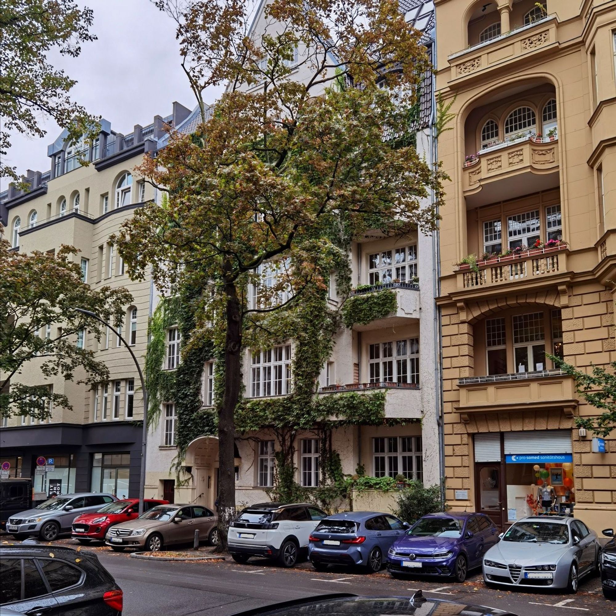 Das Foto zeigt aus einem leicht seitlichen Blickwinkel das mehrgeschossige, alte Mietshaus in der Xantener Straße 11 in Berlin-Wilmersdorf. Der Blick auf dem Bild führt über die Straße und hinweg über mehrere Pkw, die vor dem Haus geparkt sind, und vorbei an einem großen, herbstlich gefärbten Laubbaum, der die Sicht auf das Haus teilweise verdeckt. Das Gebäude hat eine helle, saubere Fassade, die über mehrere Stockwerke hinweg dicht von Efeu umrankt ist und zudem viele große Fenster und mehrere Balkone aufweist. Der Himmel in der linken oberen Bildecke ist dicht bewölkt. Das beschriebene Bauwerk befindet sich zwischen weiteren großen Mietshäusern.