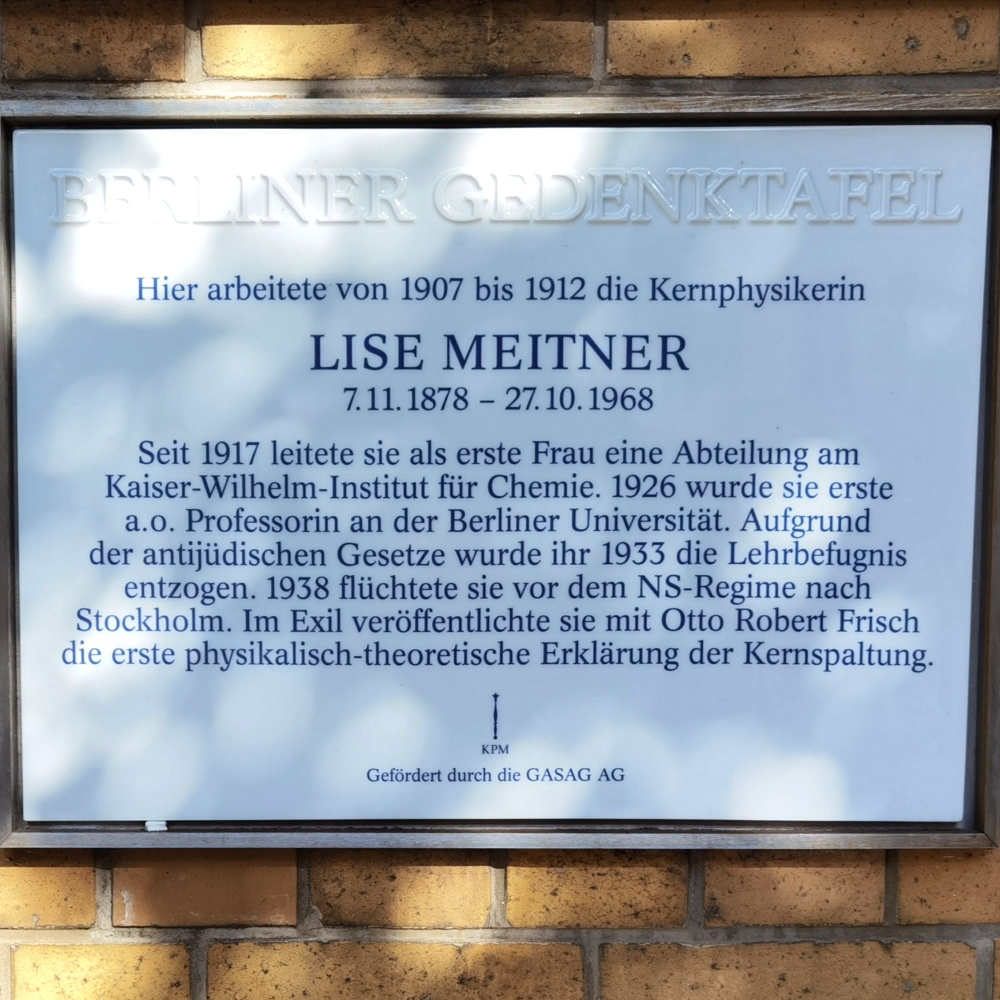 Das Foto zeigt die Gedenktafel zu Ehren von Lise Meitner in der Hessischen Straße 1 in Berlin-Mitte, darauf die folgende Inschrift: 

"Hier arbeitete von 1907 bis 1912
die Kernphysikerin

LISE MEITNER
7. 11. 1878 - 27. 10. 1968

Seit 1917 leitete sie als erste Frau eine Abteilung
am Kaiser-Wilhelm-Institut für Chemie. 1926 wurde sie erste
a.o. Professorin an der Berliner Universität. Aufgrund
der antijüdischen Gesetze wurde ihr 1933 die Lehrbefugnis
entzogen. 1938 flüchtete sie vor dem NS-Regime nach
Stockholm. Im Exil veröffentlichte sie mit Otto Robert Frisch
die erste physikalisch-theoretische Erklärung der Kernspaltung." 

Die Tafel besteht aus weißem Porzellan und ist dunkelblau beschriftet. Sie ist zudem an einer hellen, gelblichen Backsteinmauer angebracht.