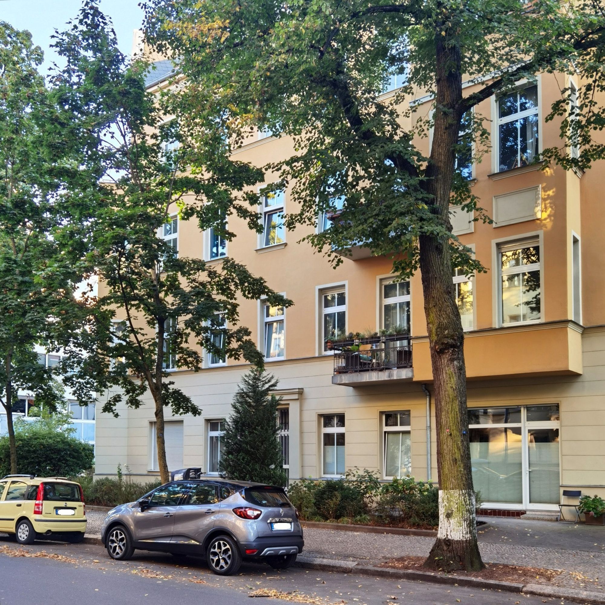 Das Foto zeigt aus einem seitlichen Blickwinkel das mehrgeschossige Mietshaus am Kronprinzendamm 20 in Berlin-Halensee. Das Haus ist ein renovierter Altbau mit heller, sauberer Fassade, die zahlreiche große Fenster, aber keine Balkone aufweist. Der Blick auf dem Bild führt vorbei an zwei parkenden Pkw vor dem Haus, das von den Kronen mehrerer üppig grünender Laubbäume teilweise verdeckt wird und zudem einen kleinen, gepflegten Vorgarten hat. Der Himmel, der in der linken oberen Bildecke zu erahnen ist, ist hellblau und unbewölkt.