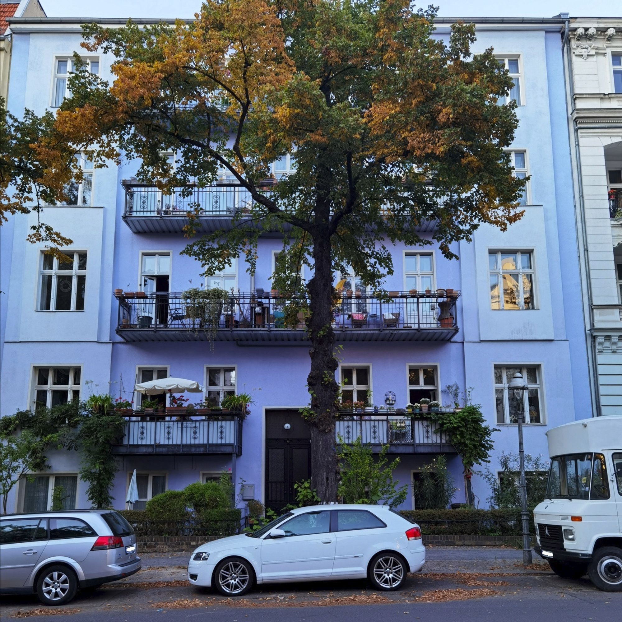 Das Foto zeigt das mehrgeschossige, alte Mietshaus am Kronprinzendamm 6 in Berlin-Halensee. Der Blick auf dem Bild führt hinweg über einen weißen Pkw, der vor dem Haus geparkt ist, und vorbei an einem großen Laubbaum mit herbstlicher Krone vor dem Haus. Die Fassade des Hauses ist sauber, hat einen hellen, blauen Farbton und weist zahlreiche, große Fenster sowie mehrere breite Balkone auf.