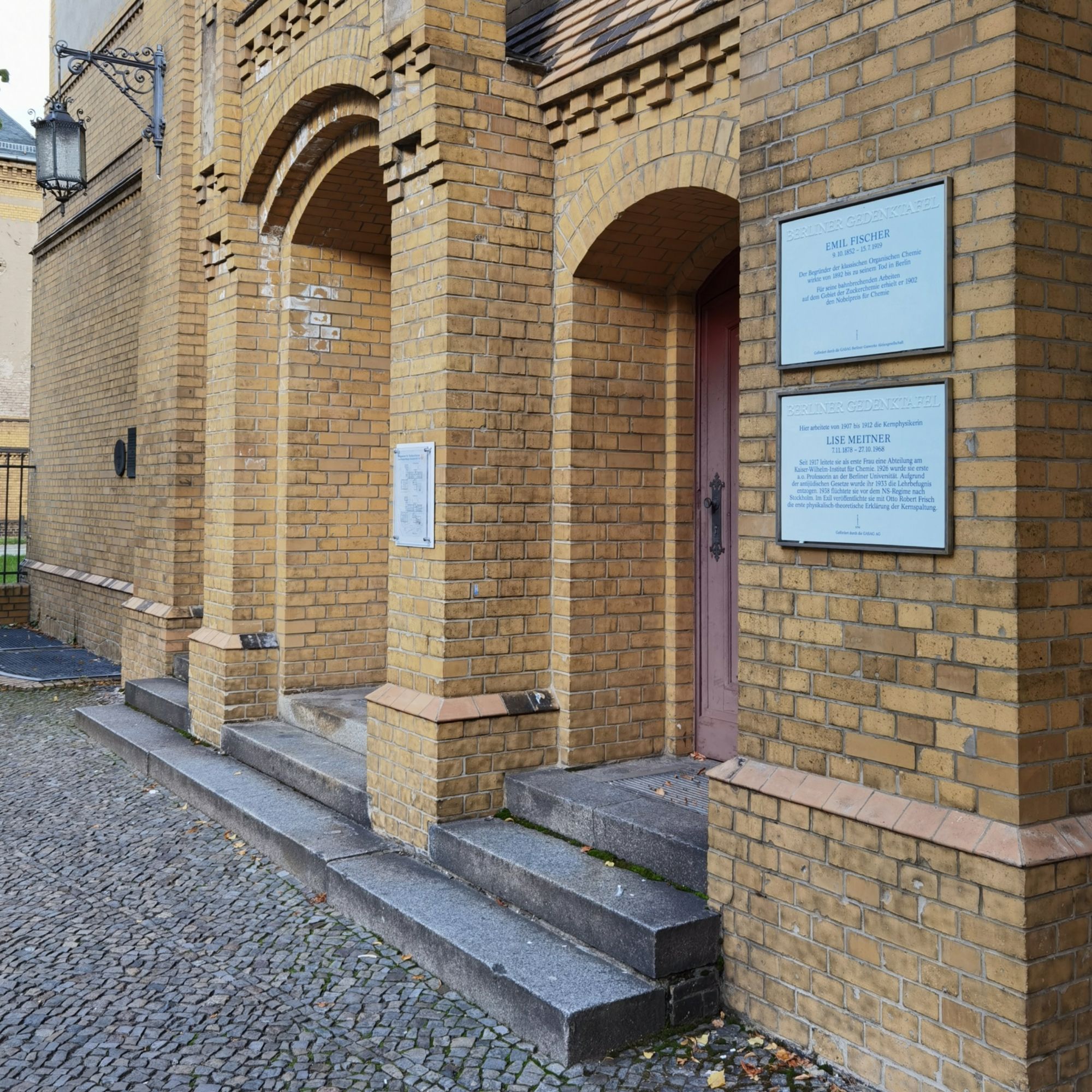 Das Foto zeigt aus einem seitlichen Blickwinkel das Portal des Institutsbaus der Humboldt-Universität zu Berlin in der Hessischen Straße 1 in Mitte. Das Mauerwerk des gesamten Eingangsbereichs besteht aus hellen, gelblichen Backsteinen, am rechten Bildrand befinden sich zwei weiße Gedenktafeln.