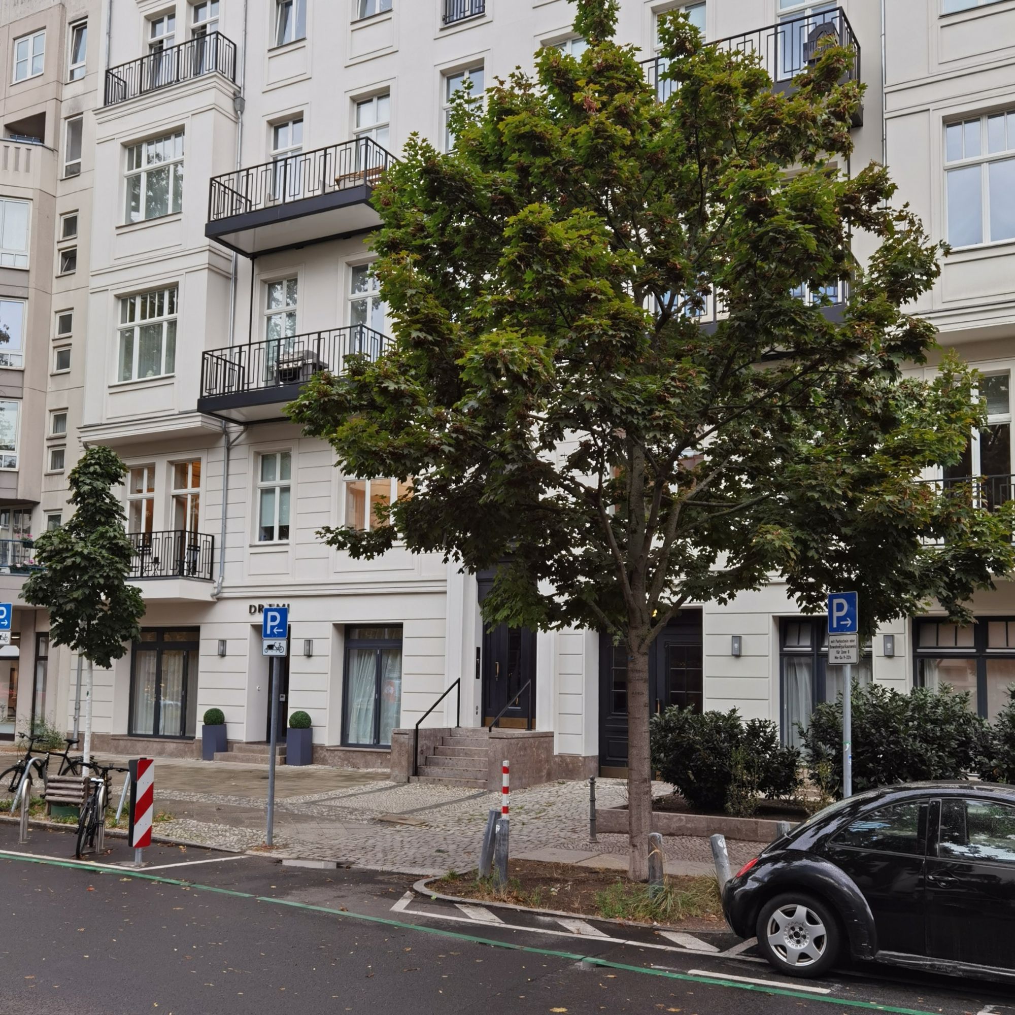 Das Foto zeigt aus einem seitlichen Blickwinkel das mehrgeschossige Mietshaus in der Fasanenstraße 65 in Berlin-Wilmersdorf. Das Haus ist ein renovierter Altbau mit heller, sauberer Fassade, zahlreichen großen Fenstern und mehreren Balkonen. Der Blick auf dem Bild führt vorbei an einem parkenden, schwarzen Pkw am rechten Bildrand und einem üppig grünenden Laubbaum vor dem Haus und führt zur verglasten Haustür, die sich im unteren Bildbereich exakt in der Bildmitte befindet.