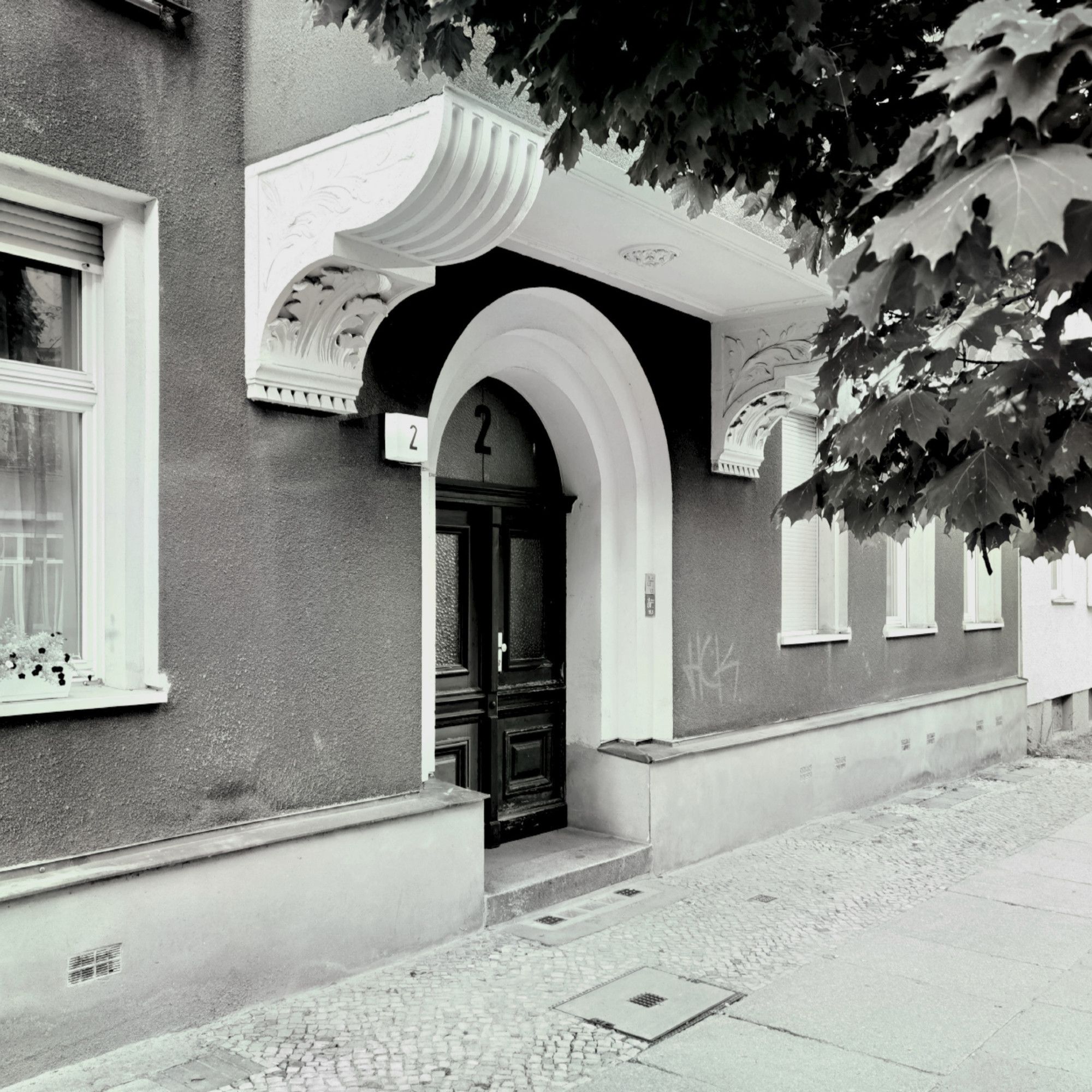 Das schwarz-weiße Foto zeigt aus einem seitlichen Blickwinkel die hölzerne, verglaste Haustür und den Eingangsbereich zum mehrgeschossigen Mietshaus in der Lützenstraße 2 in Berlin-Halensee. Die Fassade des Hauses ist dunkel und sauber und weist mehrere große Fenster auf, direkt über der Haustür ist ein prachtvoller Balkon zu erahnen. Die Blätter eines üppig grünenden Laubbaumes ragen in der rechten oberen Bildecke in das Blickfeld.