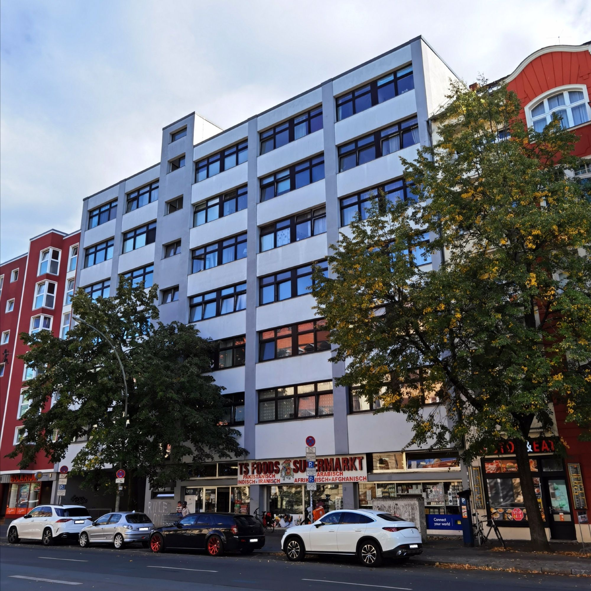 Das Foto zeigt aus einem leicht seitlichen Blickwinkel das moderne, mehrgeschossige Mietshaus in Alt-Moabit 74 in Berlin-Moabit. Der Blick auf dem Bild führt hinweg über die Straße und mehrere parkende Pkw bzw. hindurch zwischen zwei großen, herbstlich gefärbten Laubbäumen. Das Haus ist ein beinahe würfelförmiger Neubau mit heller Fassade, einem flachen Dach und sehr zahlreichen Fenstern. Die Schaufenster eines Supermarktes bestimmen das Erdgeschoss des Hauses, das sich direkt zwischen weiteren Wohnhäusern befindet. Der Himmel am oberen Bildrand ist hellblau und leicht bewölkt.