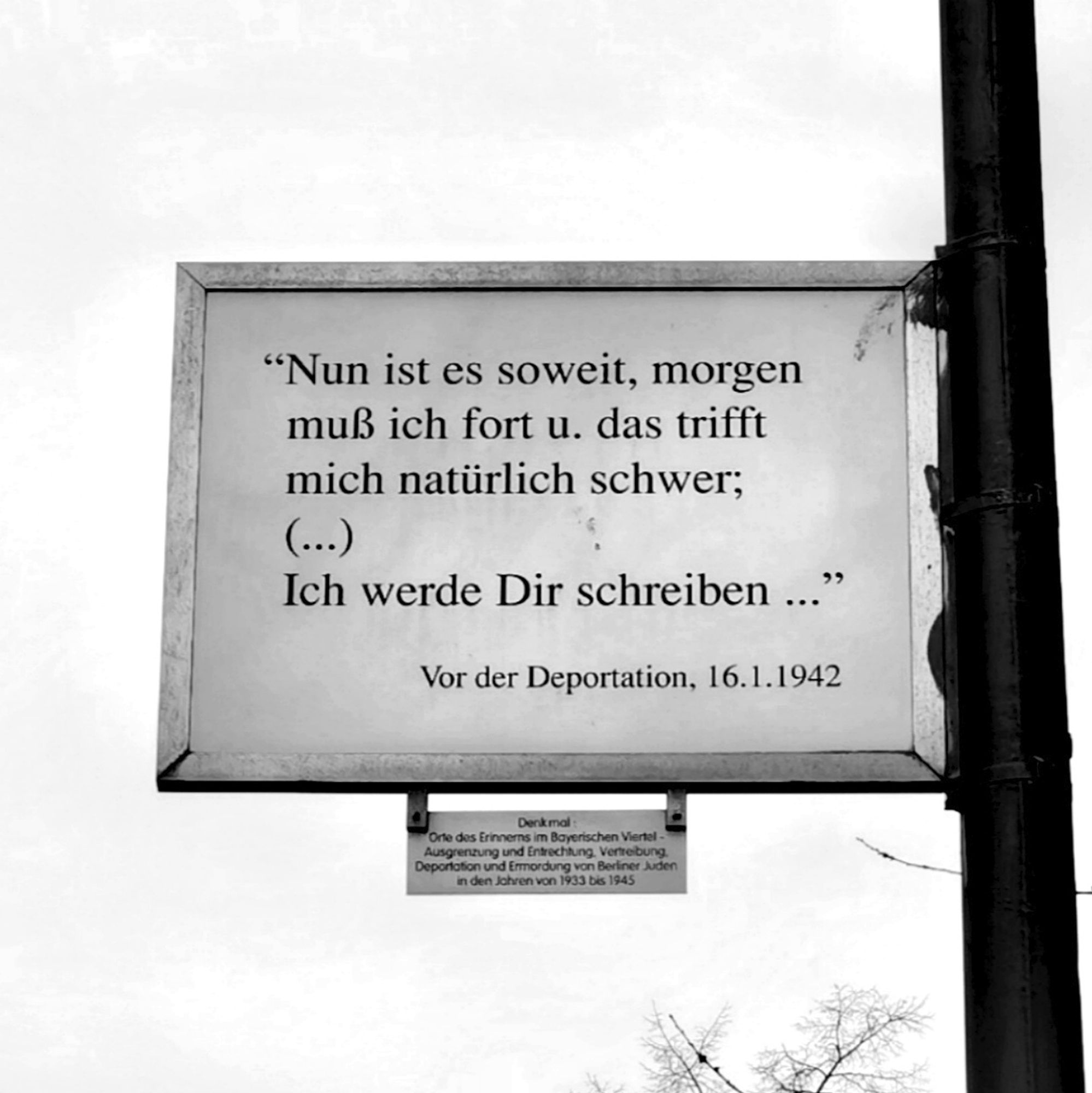 Das schwarz-weiße Foto zeigt eine Gedenktafel im Bayerischen Platz in Berlin-Schöneberg, darauf die folgende Inschrift: "'Nun ist es soweit, morgen muß ich fort u. das trifft mich natürlich schwer; (...) Ich werde Dir schreiben...' Vor der Deportation, 16. 1. 1942". Die Tafel befindet sich an einem Laternenpfahl, der am rechten Bildrand in das Blickfeld ragt. Das Mahnzeichen hebt sich von einem dicht bewölkten, hellen Himmel im Bildhintergrund ab, am unteren Bildrand ist der oberste Teil der kahlen Krone eines Laubbaumes zu erahnen.