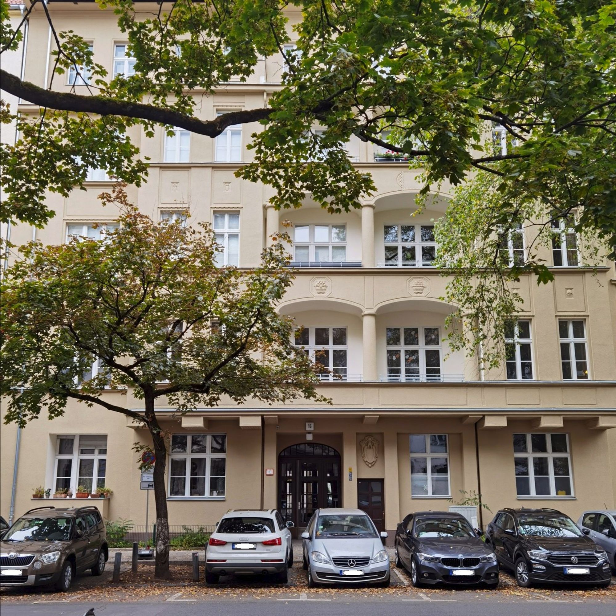 Das Foto zeigt das mehrgeschossige Mietshaus in der Xantener Straße 16 in Berlin-Wilmersdorf. Das Haus ist ein renovierter Altbau mit beiger, sauberer Fassade, sehr zahlreichen Fenstern und mehreren Balkonen. Der Blick auf dem Bild führt über die Straße und hinweg über mehrere parkende Pkw zur hölzernen, verglasten Haustür, die sich im unteren Bildbereich in der Bildmitte befindet. Die üppig grünenden Zweige eines großen Laubbaumes ragen am oberen Bildrand in das Blickfeld, ein weiterer Laubbaum befindet sich am linken Bildrand zwischen den parkenden Autos.