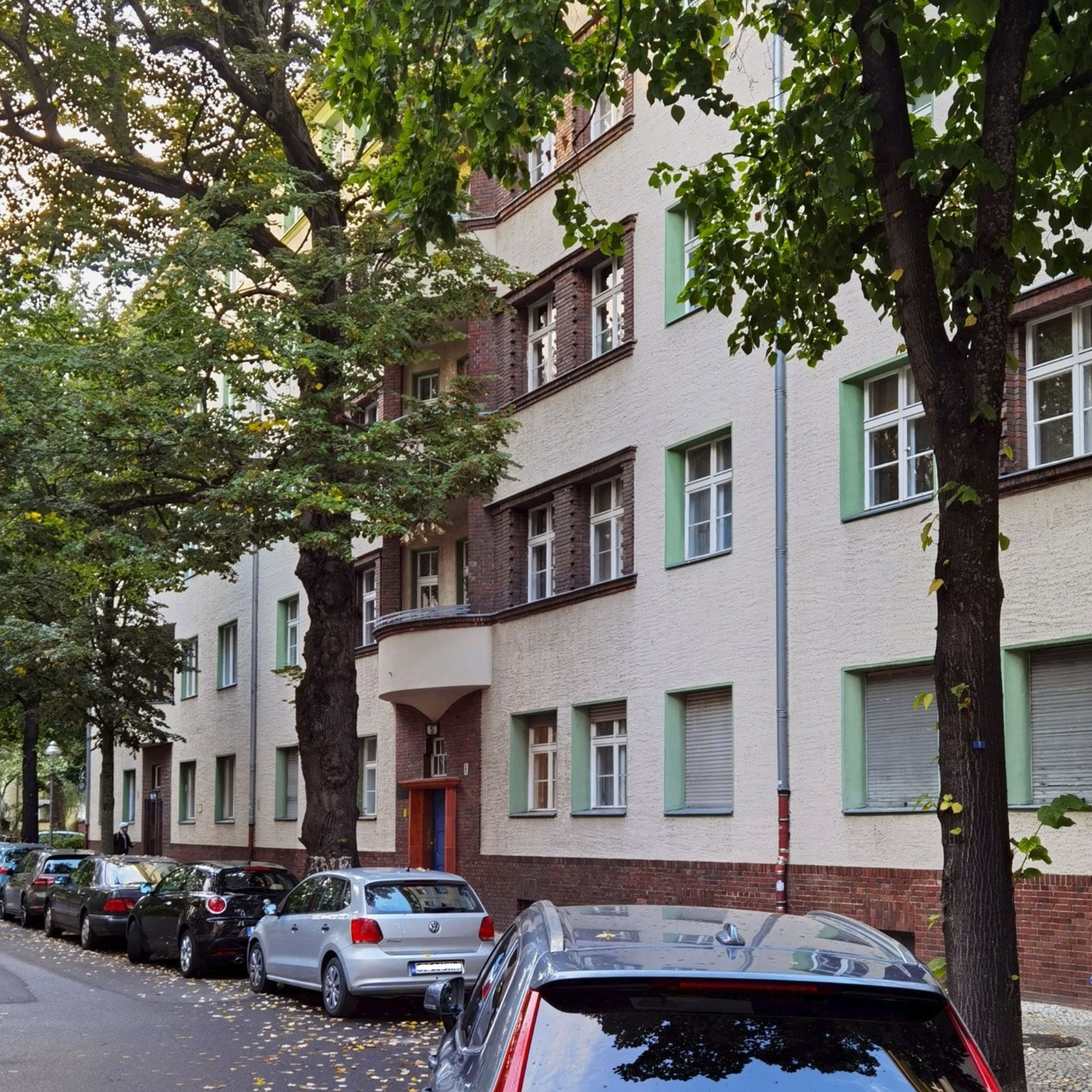 Das Foto zeigt aus einem seitlichen Blickwinkel das mehrgeschossige, alte Mietshaus in der Wittelsbacherstraße 5 in Berlin-Wilmersdorf. Der Blick auf dem Bild führt hinweg über mehrere parkende Pkw und vorbei an mehreren üppig grünenden Laubbäumen auf dem Gehweg vor dem Haus. Der Bau selbst hat eine helle, saubere Fassade mit zahlreichen Fenstern, aber ohne Balkone.