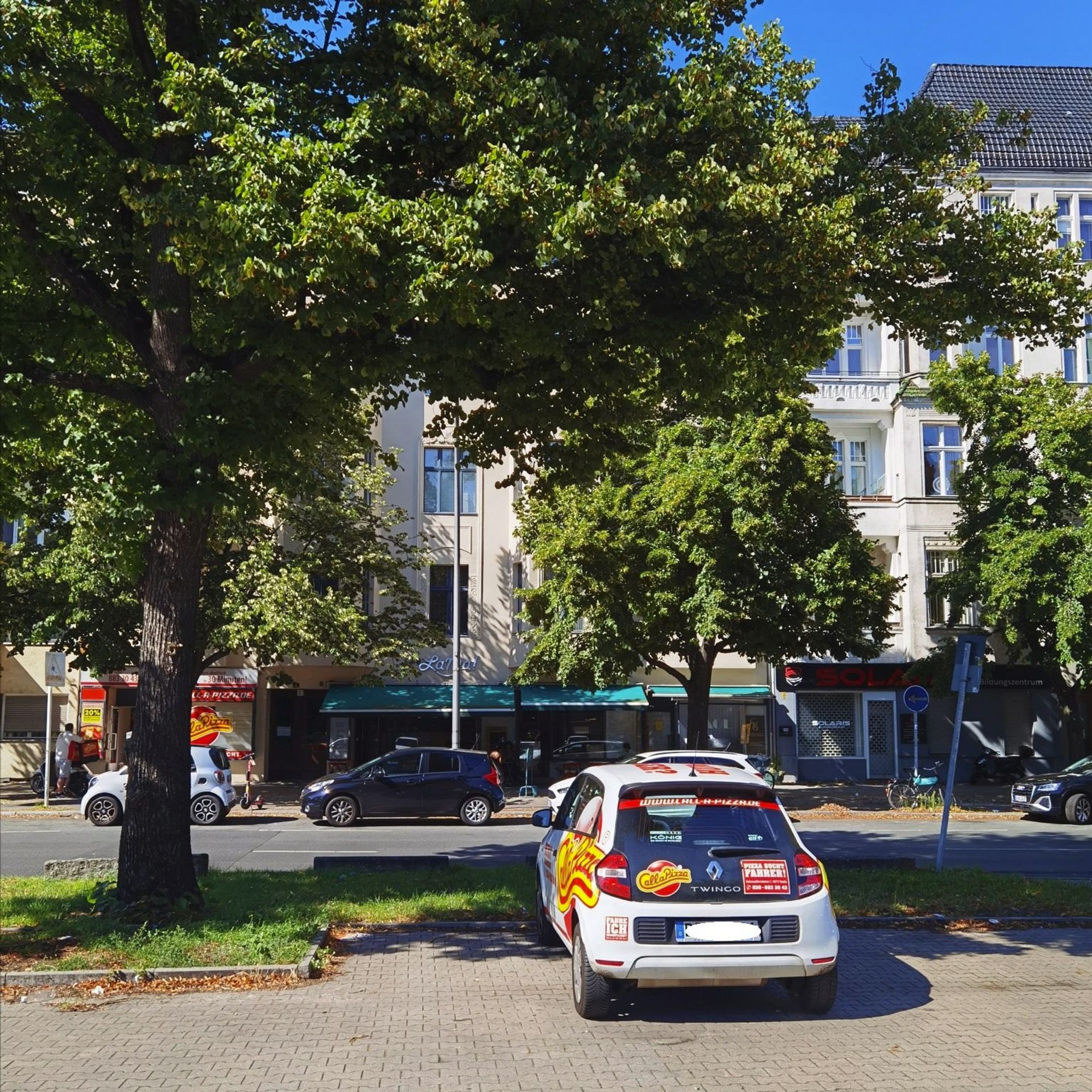 Das Foto zeigt einen Blick auf das mehrgeschossige Mietshaus am Hohenzollerndamm 7 in Berlin-Wilmersdorf. Der Blick auf dem Bild führt von einer Parkfläche an der besagten Straße hinweg über das Heck eines weißen Pkw, das mit großflächigen Werbeaufklebern für einen Pizzalieferdienst in roter und in gelber Farbe auffällt. Die Krone des großen, üppig grünenden Laubbaumes am linken Bildrand verdeckt die oberen Stockwerke des Hauses, vor dem weitere Bäume und zudem mehrere geparkte Pkw stehen. Das Haus hat eine helle, saubere Fassade mit zahlreichen großen Fenstern, in seinem Erdgeschoss befinden sich ein Restaurant in der Bildmitte und ein Pizzalieferdienst am linken Bildrand. Der Himmel, der am oberen Bildrand zu erahnen ist, ist tiefblau und unbewölkt.