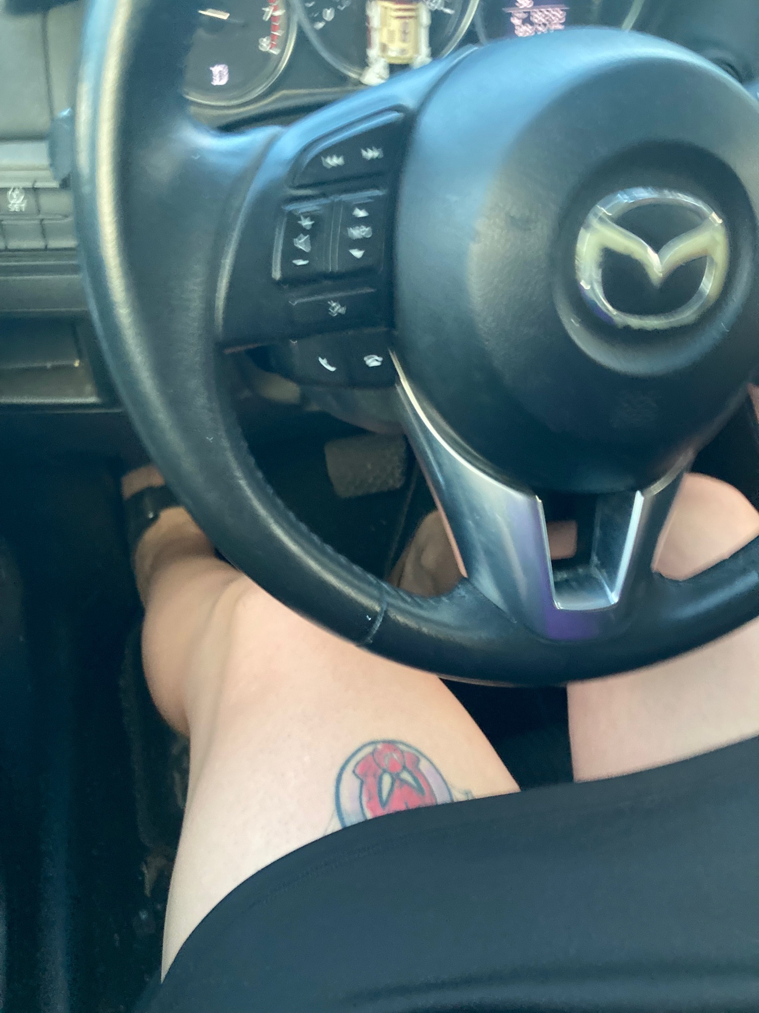 Driver’s point of view of Lauren driving. We see her legs extending out of a black skirt that hits midthigh. A tattoo is showing on the top of her left thigh.