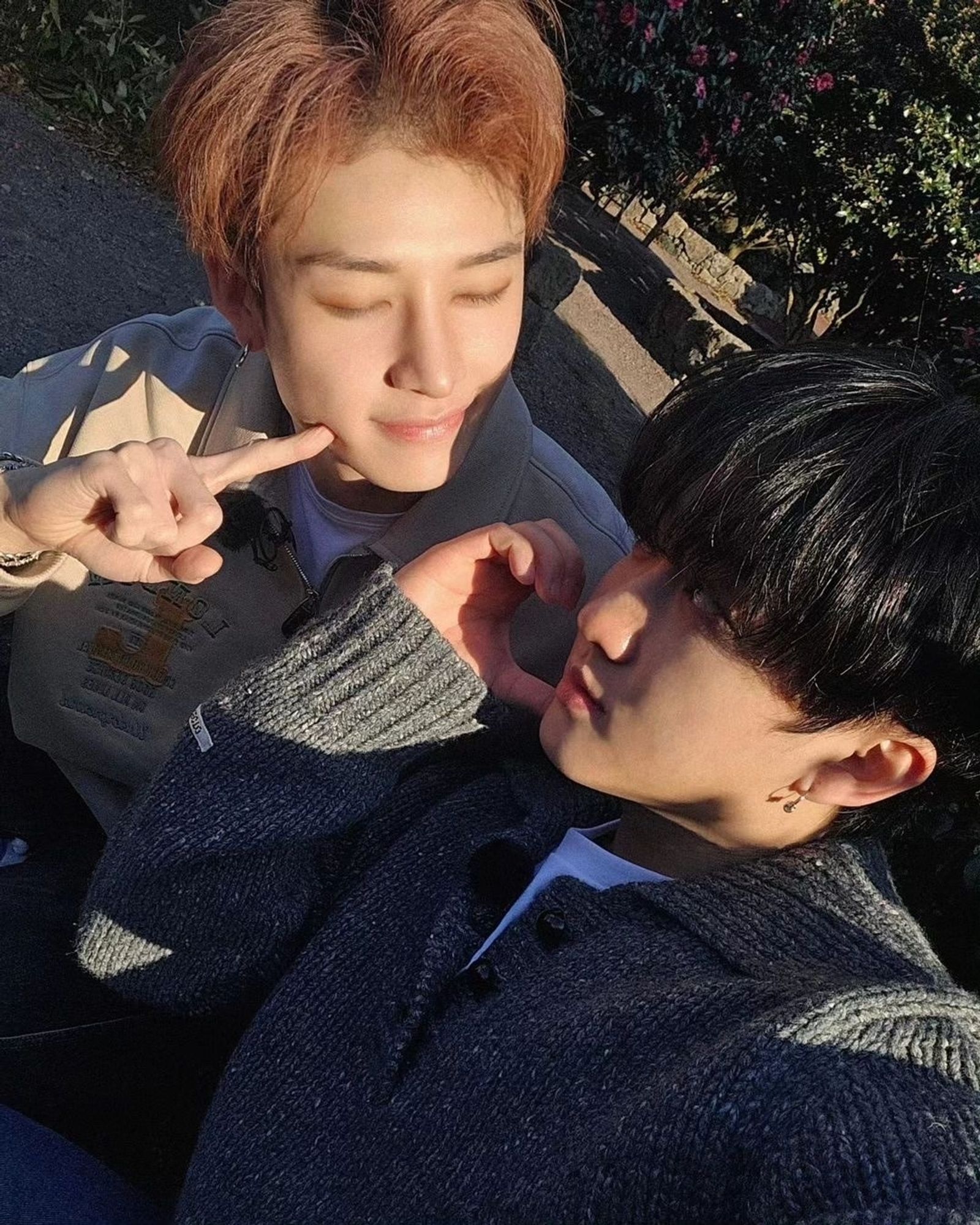 This is a selfie of Changbin with Bang Chan. Changbin is wearing a gray sweater and Bang Chan is wearing a beige hoodie. The angle of the photo is from above. Changbin is taking the photo. He is looking at Bang Chan and is making half a heart with his right hand next to his face. Bang Chan is facing Changbin (on his left) with his eyes closed and his right index finger is on his right cheek. In the background there are trees