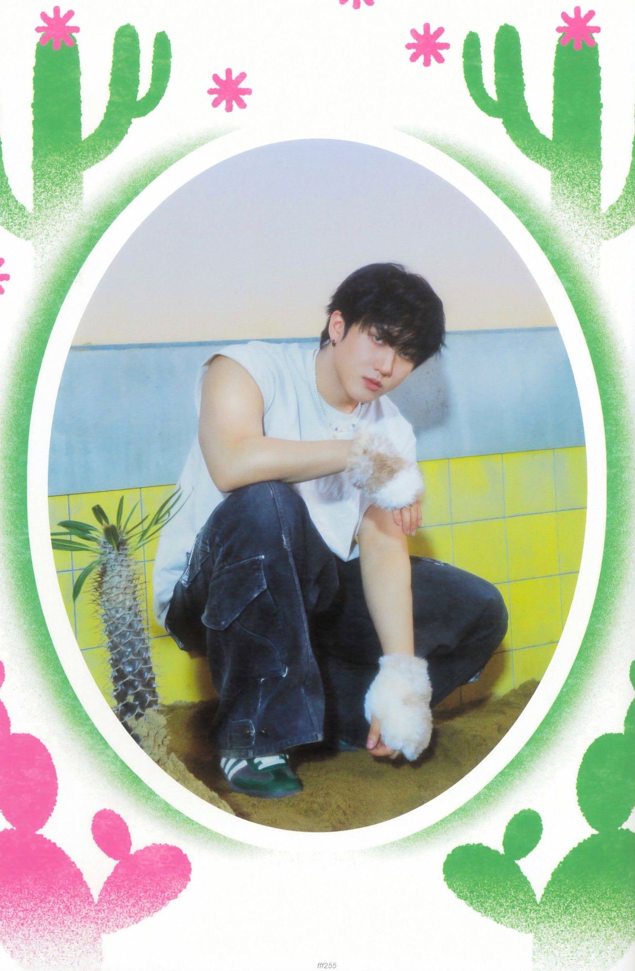 Changbin is dressed in a white t-shirt and dark jeans, and white plush gloves. He is crouched inside an empty pool with sand and yellow tiles. He is looking at the camera and there are several cacti in the image  